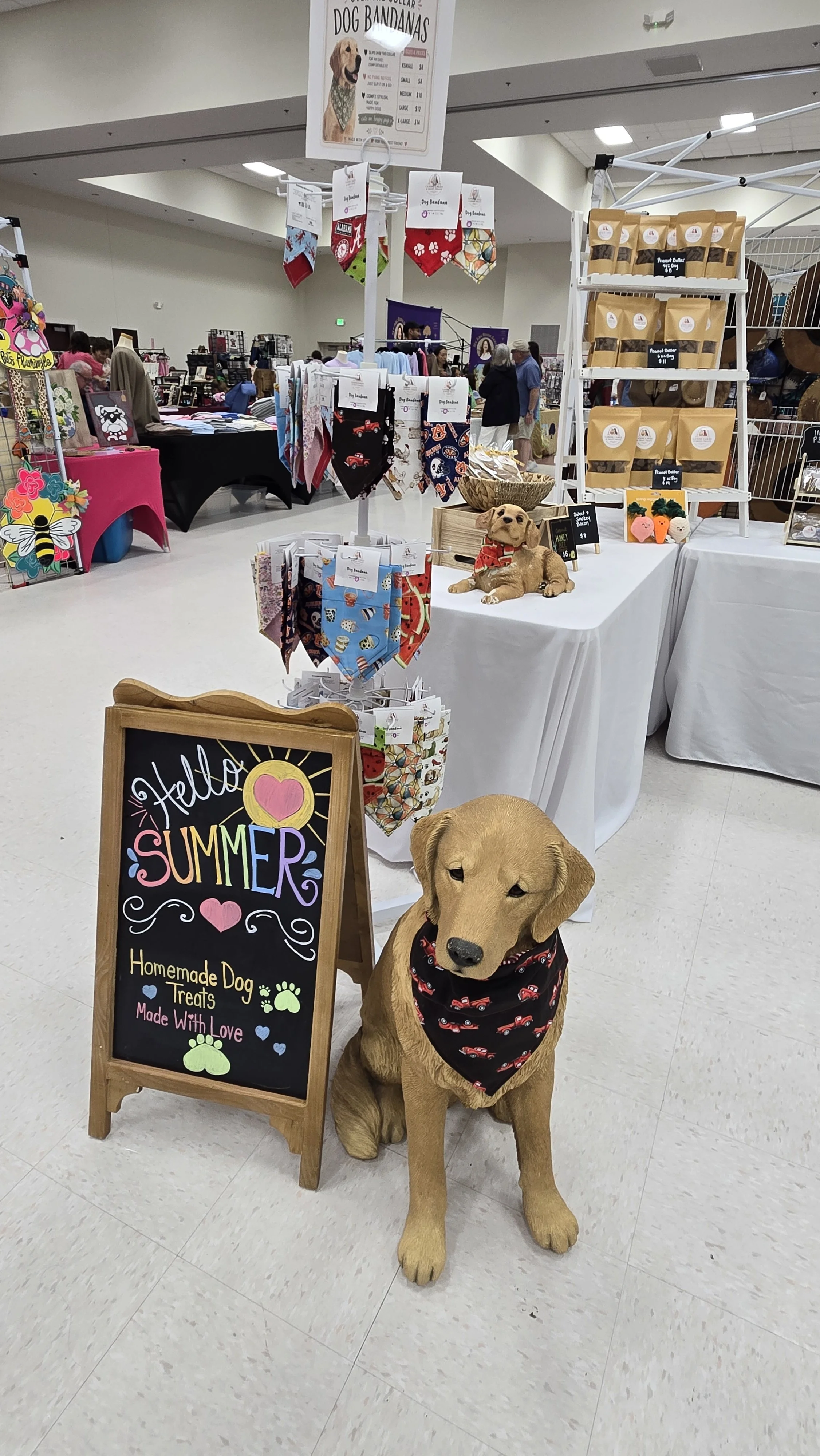 A yellow stuffed dog wearing a bandana sits next to a chalkboard sign that reads 'Hello Summer, Homemade Dog Treats Made With Love' in a shopping mall. Behind the dog, there is a display table with various pet accessories and treats, and other vendor