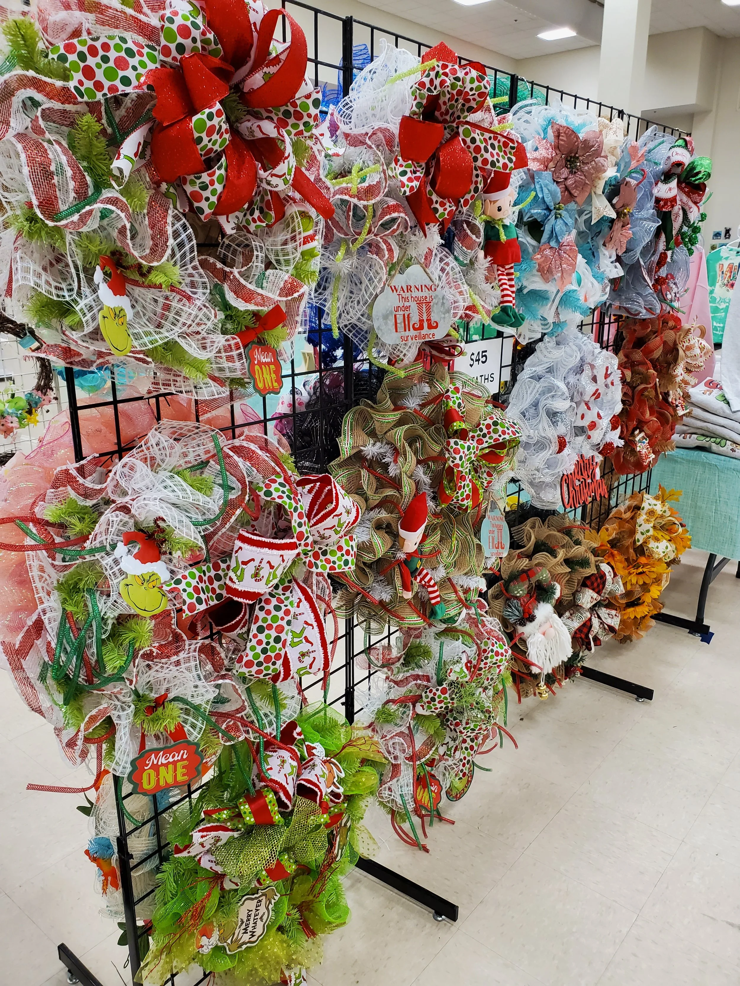 Display of various Christmas wreaths and decorations on a black metal stand in a store, featuring bows, ribbons, and holiday ornaments in red, white, green, and other festive colors.