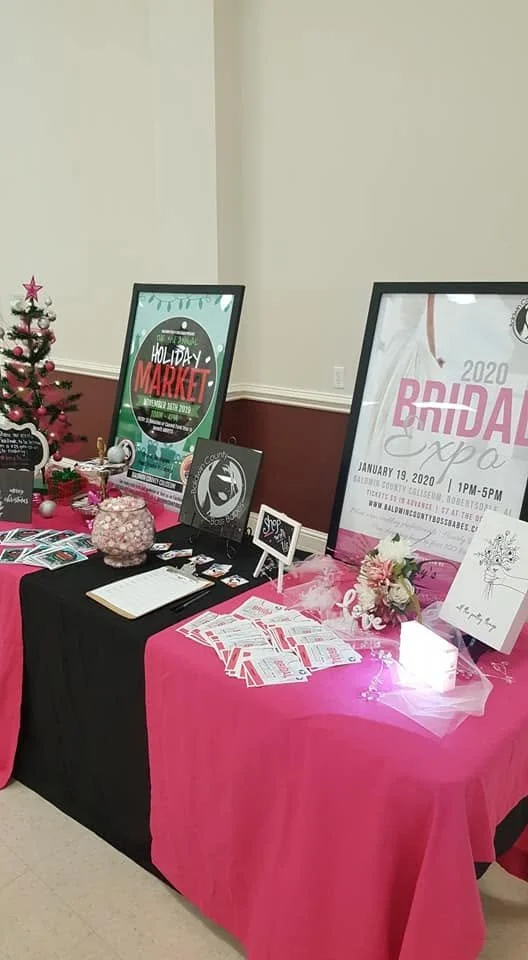 Information booth at a bridal expo with pink and black tablecloths, brochures, flowers, and family-friendly decorations, including a small Christmas tree.