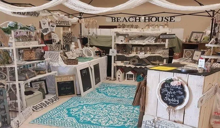 A vintage-style boutique shop with various home decor and gift items, including picture frames, signs, and decorative accessories, set up in a cozy, rustic display at a market or craft fair.