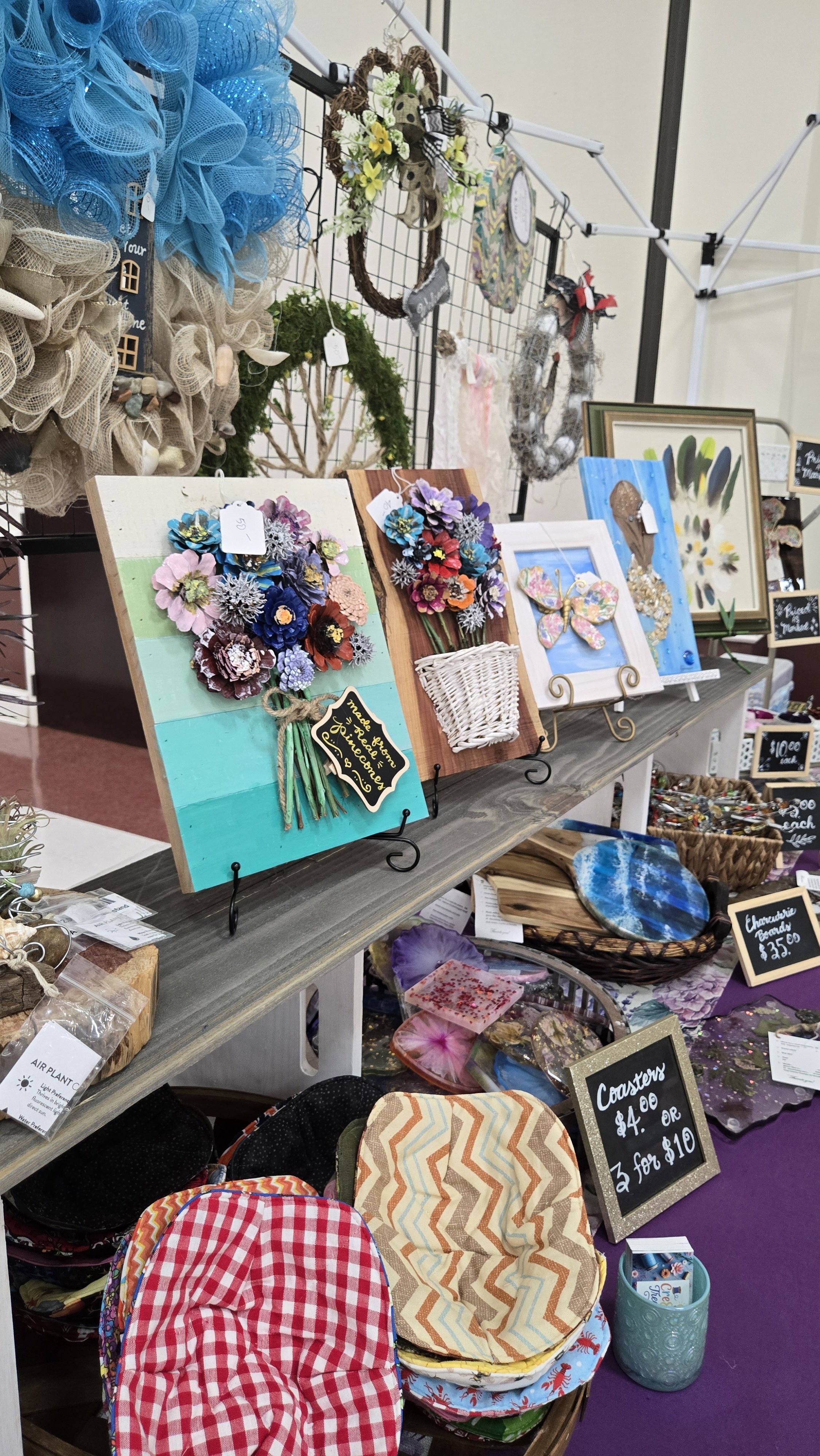 Display table at a craft fair with handmade wall decorations, framed art pieces, and cloth pot holders in various patterns and colors.