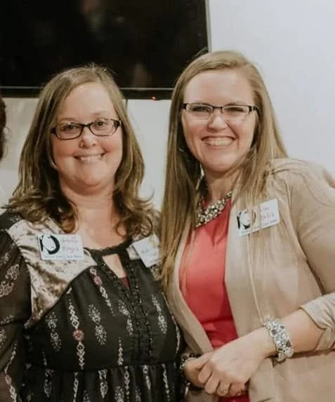 Two women smiling and shaking hands at an indoor event, both wearing glasses and name tags.