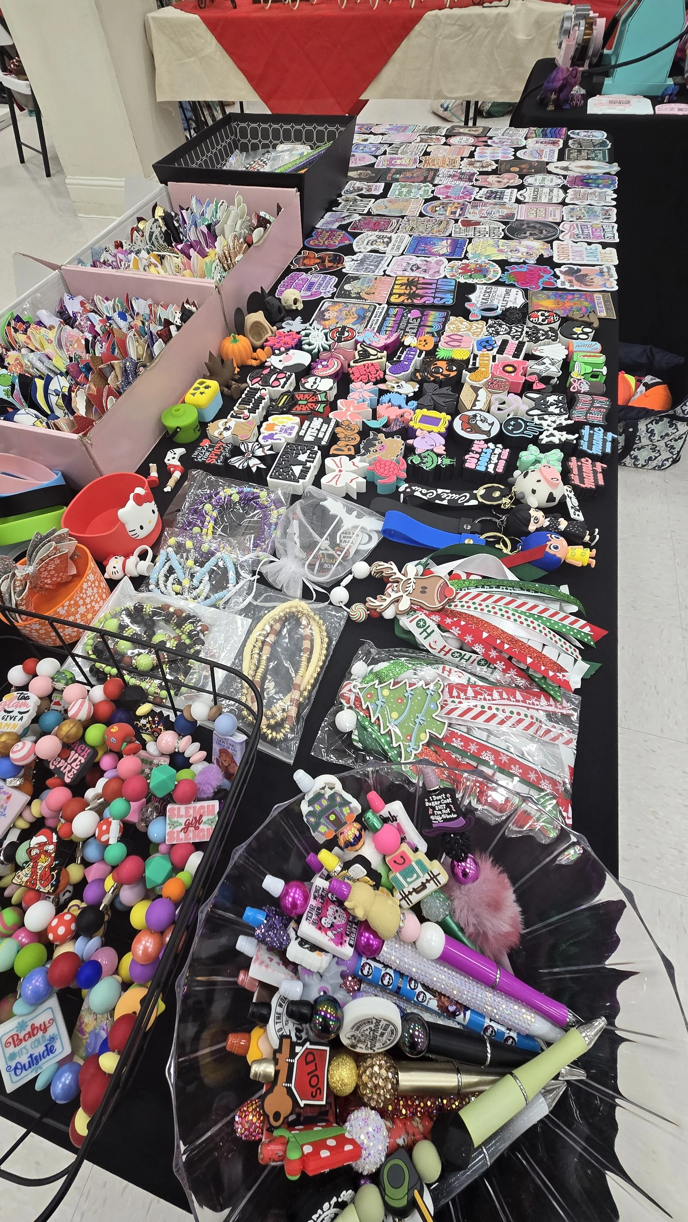 A table at a craft fair or market displaying a variety of small accessories, toys, keychains, and decorations, including holiday-themed items, with more items in boxes and on tables in the background.