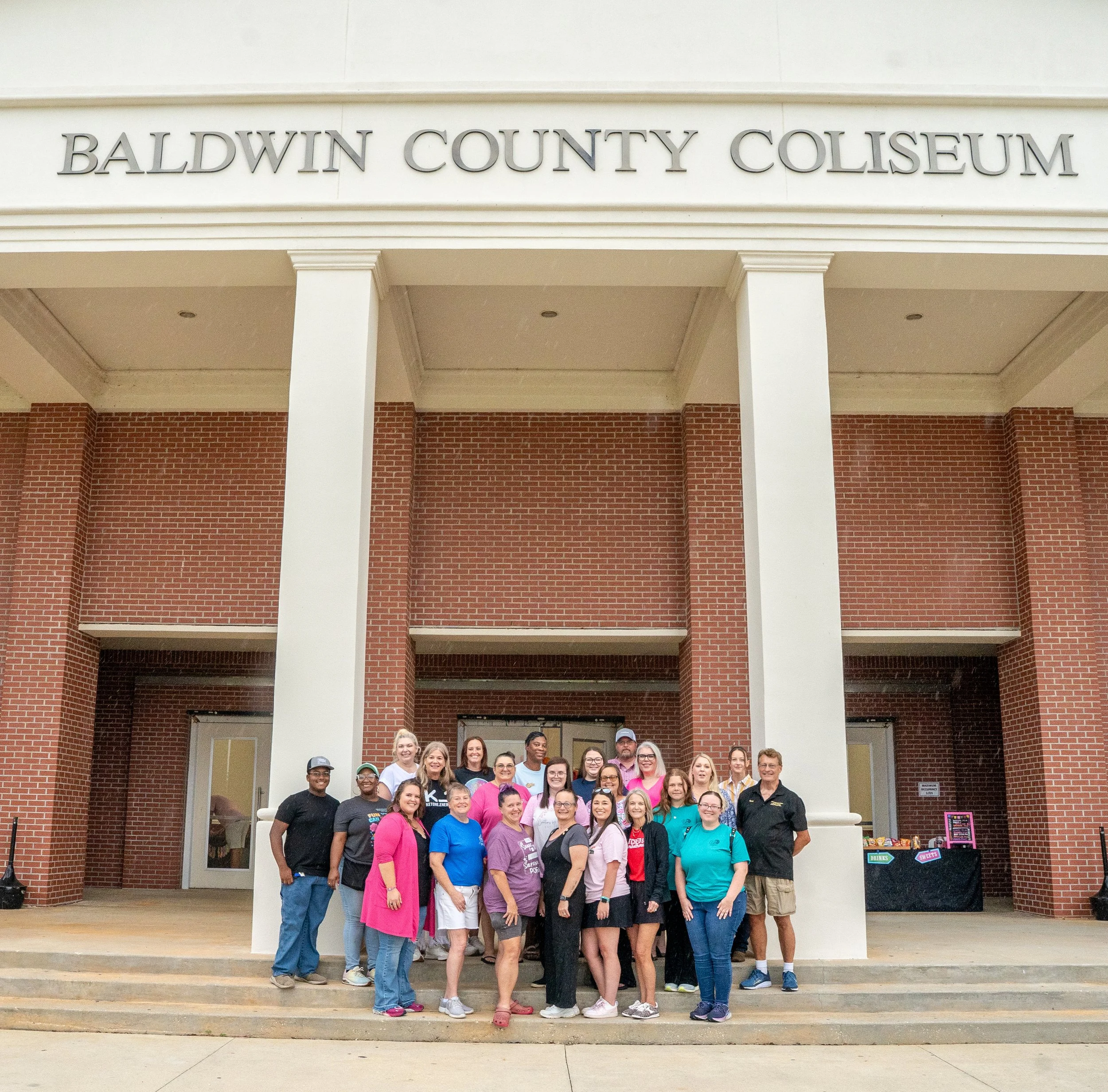 Group of people standing on the steps outside the Baldwin County Coliseum, smiling and posing for a photo