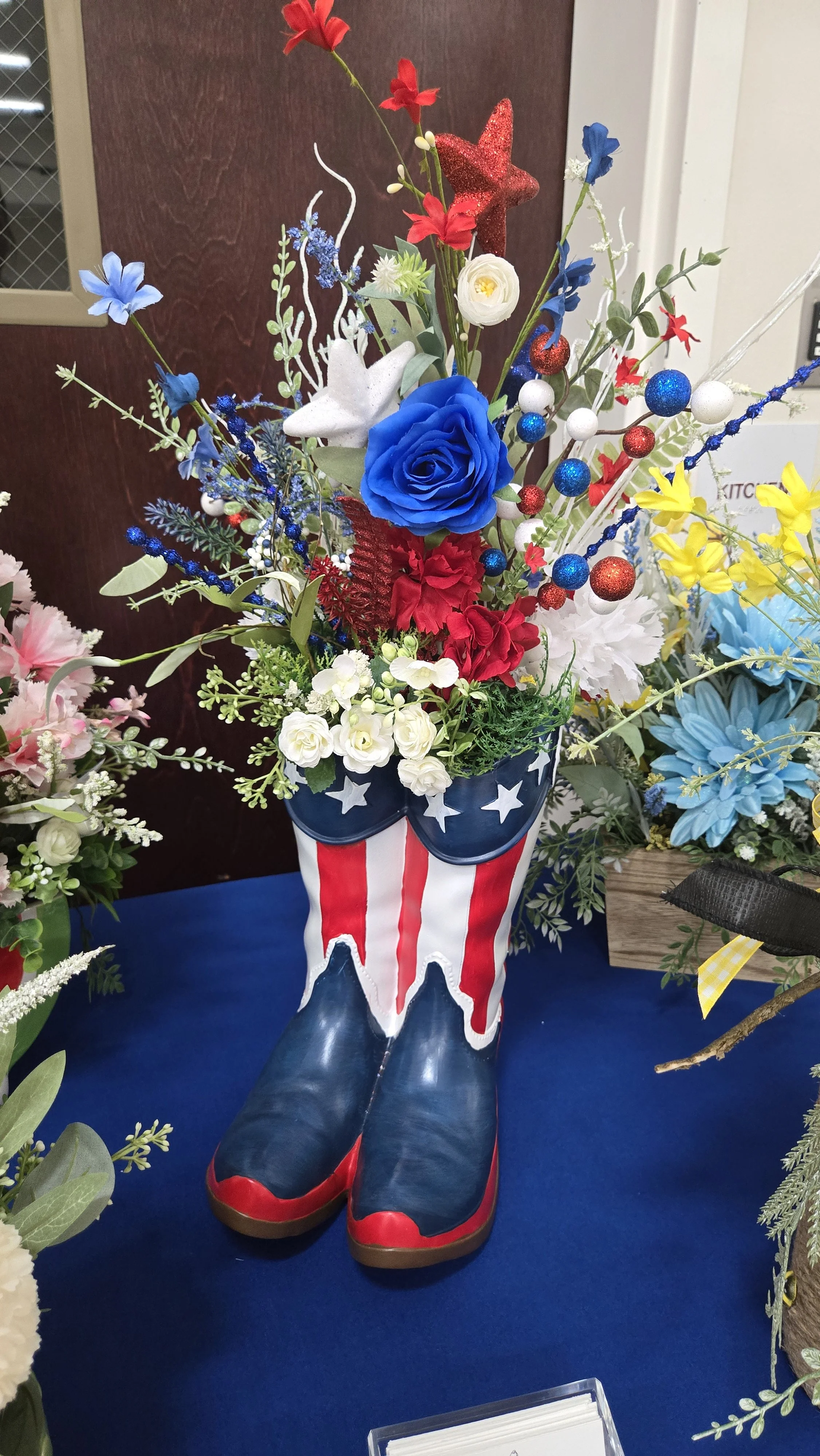 A decorative arrangement of artificial flowers in a patriotic-themed cowboy boot vase, featuring blue, red, white, and yellow flowers, with a star and stripe pattern on the boot.