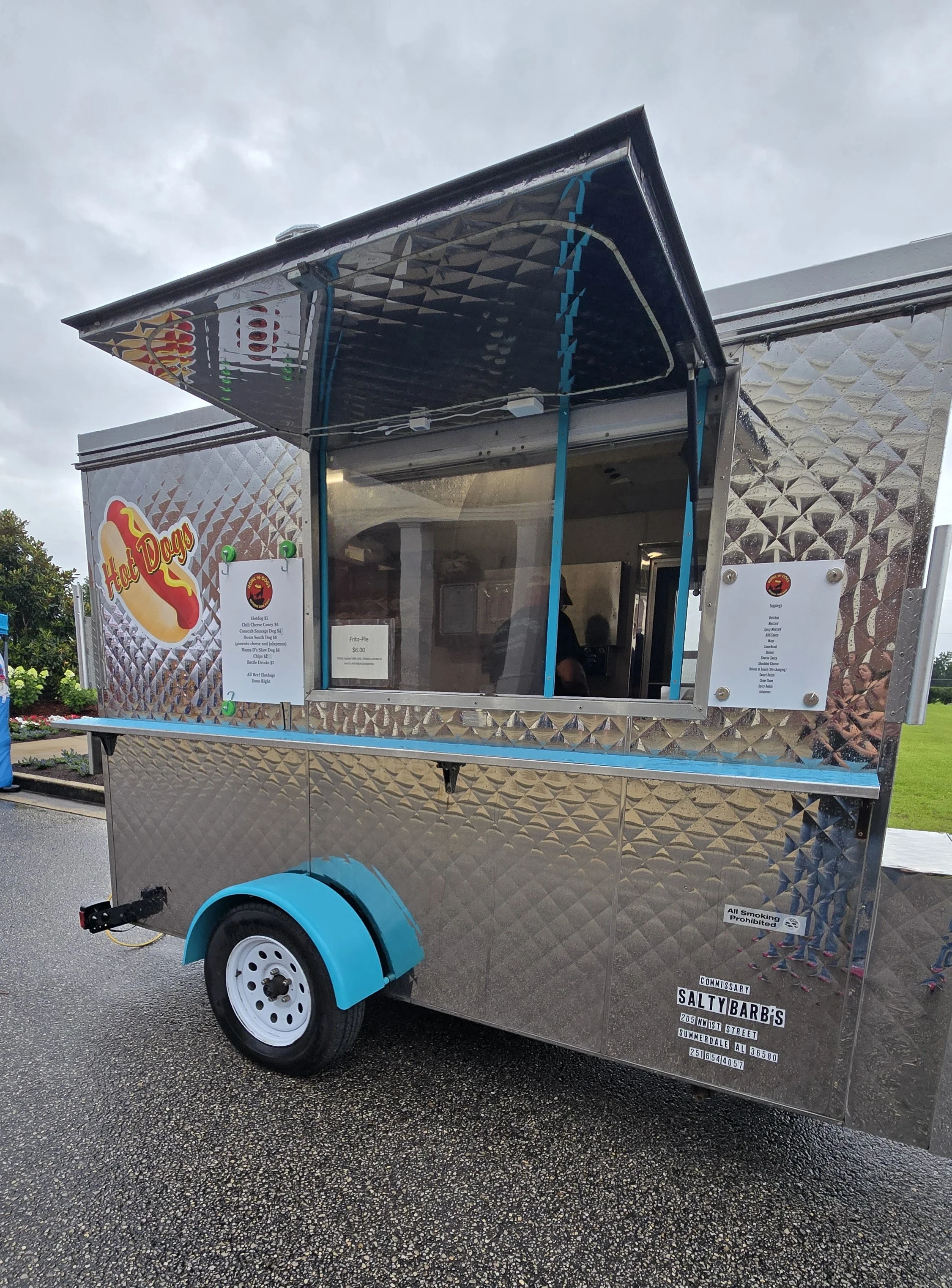 A food truck with a shiny, diamond-patterned metal exterior, featuring a sign that says 'Hot Dogs' and a menu listing various hot dog options. The truck has blue accents and is parked on a wet street.