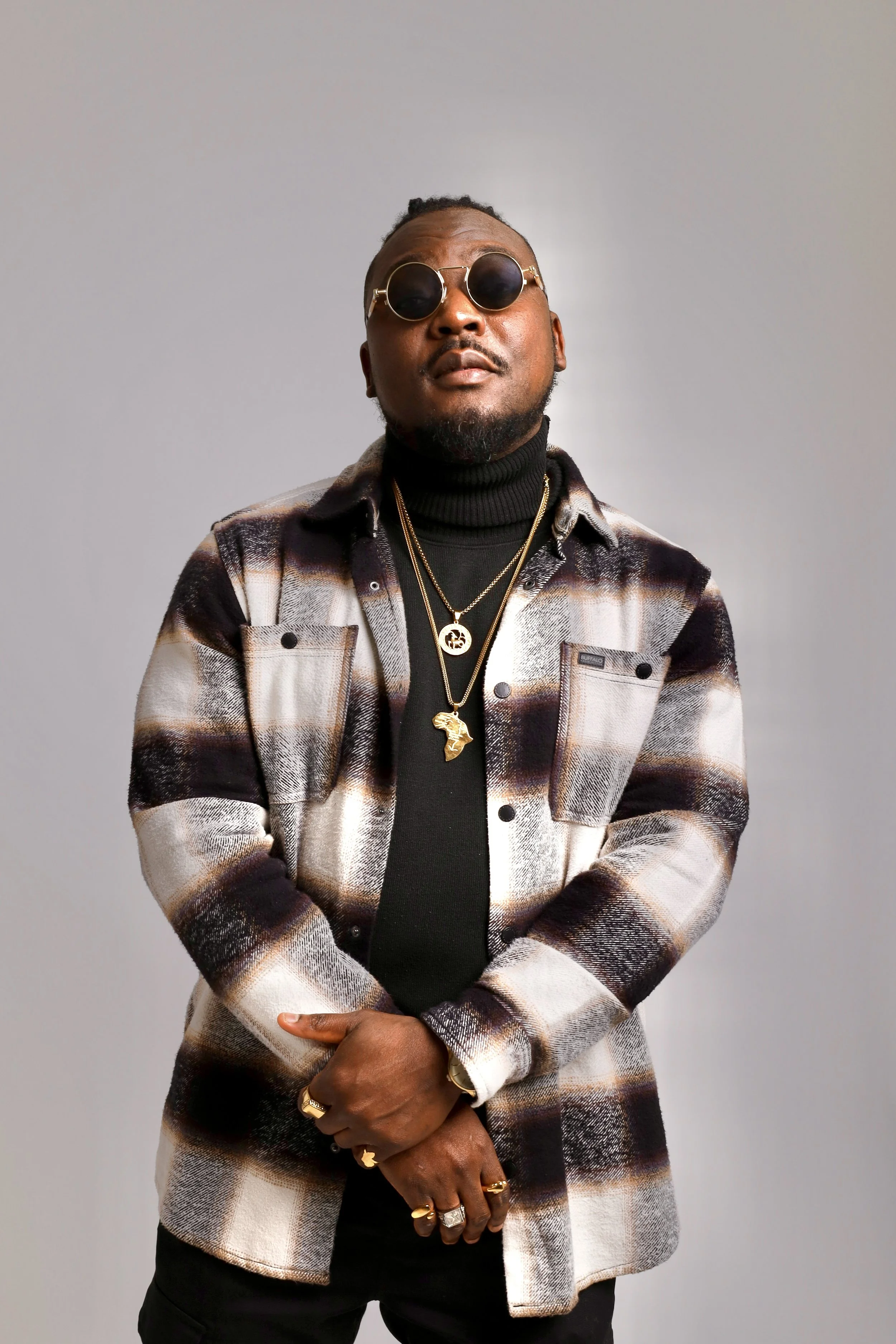 Man wearing sunglasses, black turtleneck, plaid jacket, and gold jewelry standing against a gray background.