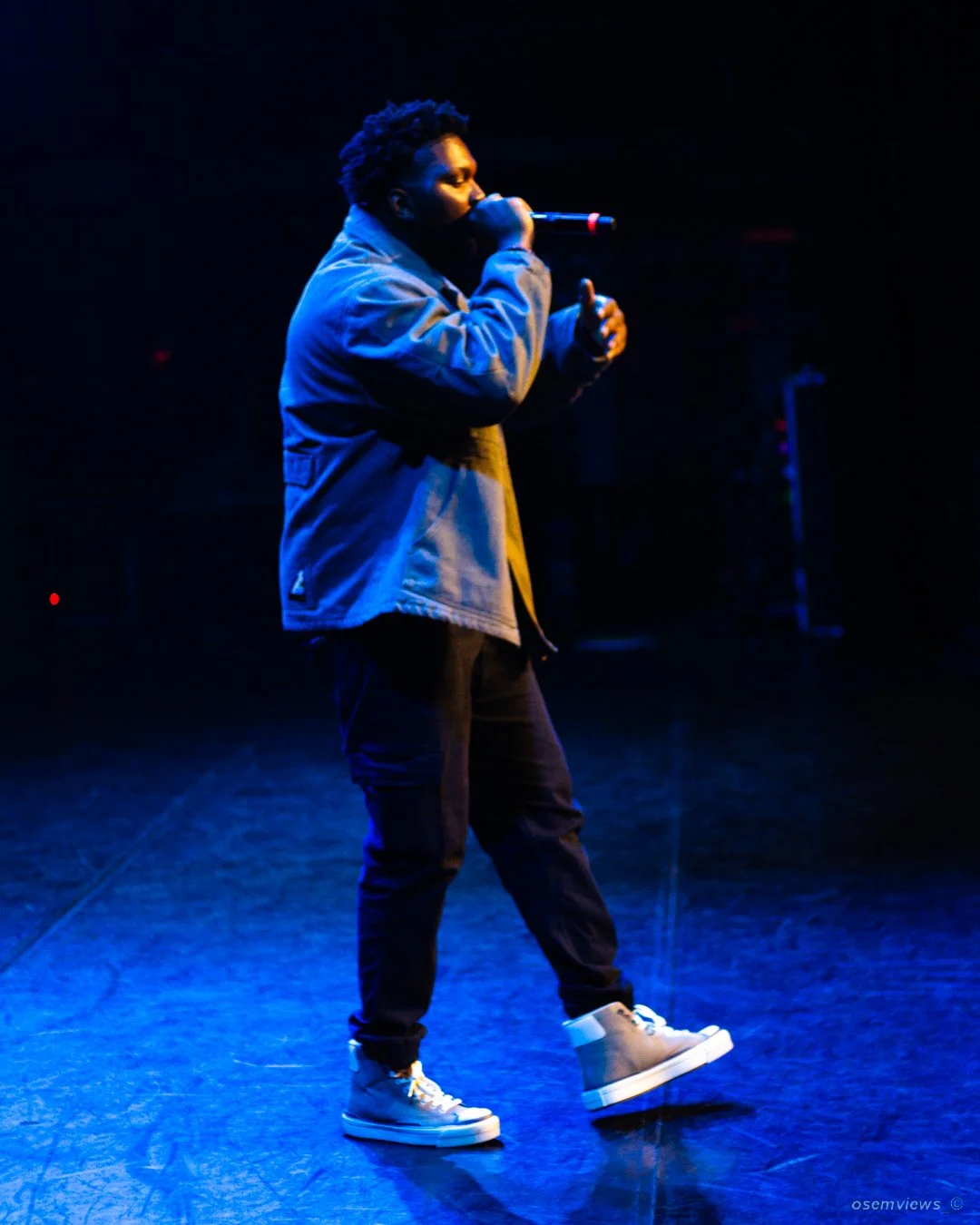 A young male rapper performing on stage, holding a microphone close to his mouth, wearing a gray jacket, black pants, and white sneakers, illuminated by blue stage lights.