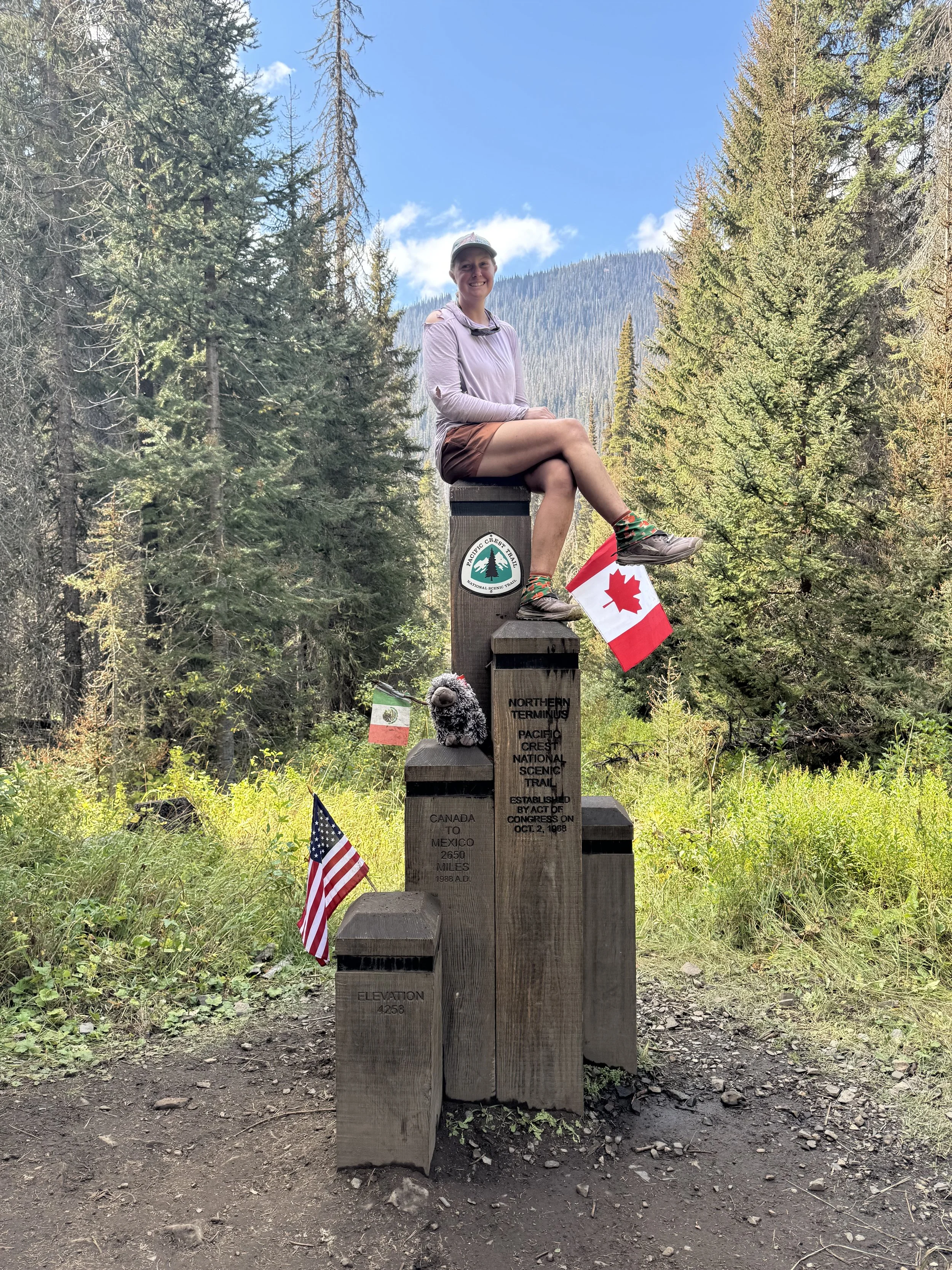 A woman, Porcupine, sits on the northern terminus monument for the Pacific Crest Trail in a forest with tall trees and mountains in the background. The monument includes flags from the United States, Canada, and Mexico and stuffed animal porcupine.