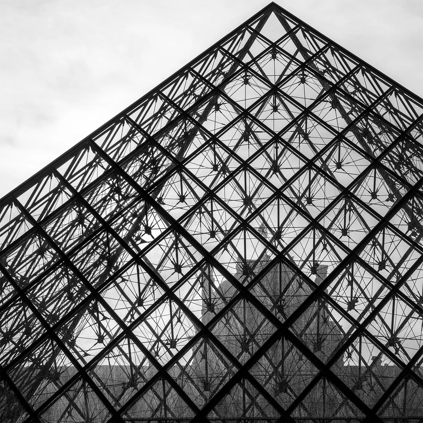 Close-up of a modern glass building with a geometric pattern, showing reflections of a bridge and trees in its mirrored surface in black and white.