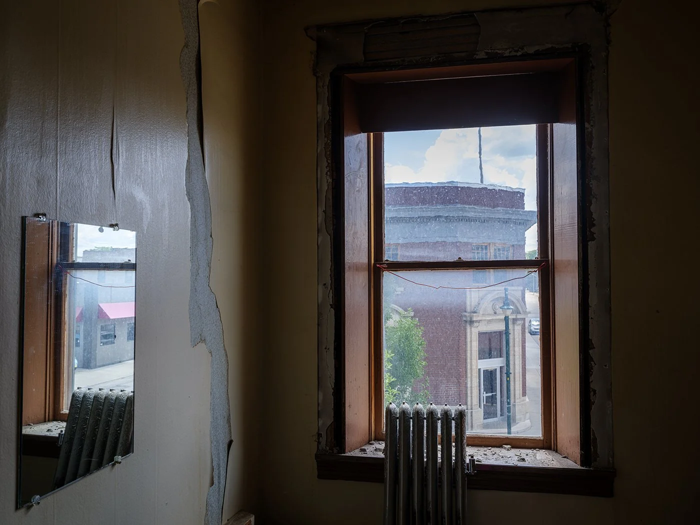 Interior view of a room under renovation showing a large window with a view of older brick buildings outside. The room has peeling paint and a damaged wall, with a cracked mirror on the left and a radiator below the window.