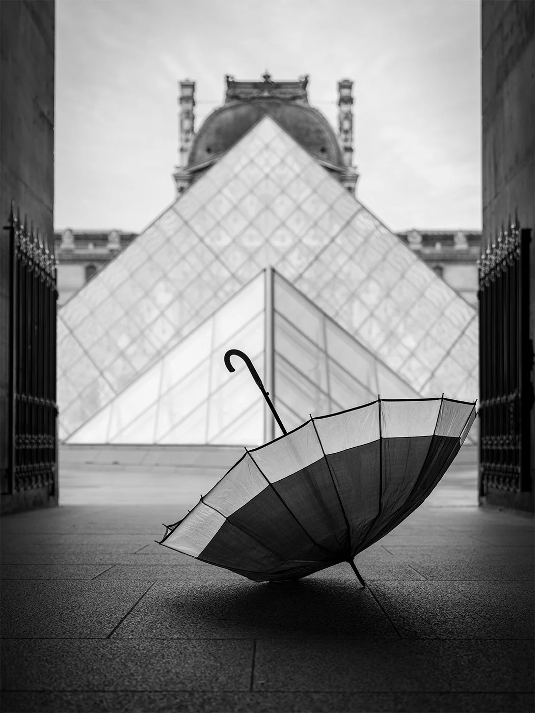 Black and white photo of an open umbrella on the ground with the Louvre Pyramid and the Louvre Museum in the background.