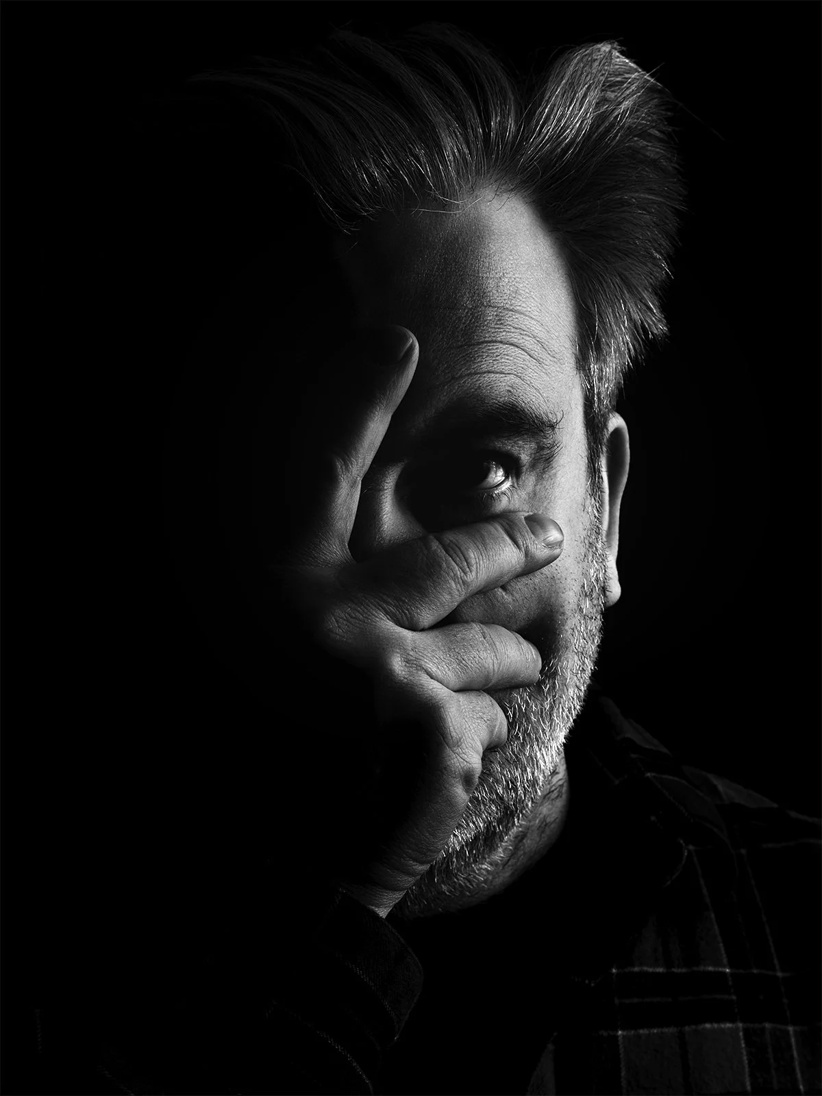 Black and white portrait of a man with gray hair and beard, covering part of his face with his hand, with one eye visible.