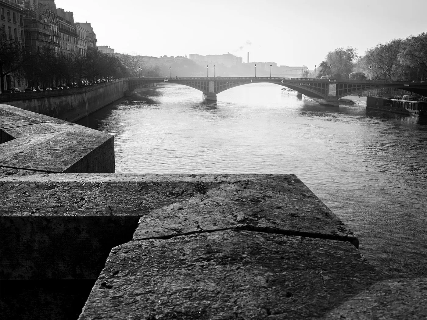 Black and white photo of a river with a bridge in the distance, stone ledge in the foreground, leafless trees along the river, and buildings on the left side.