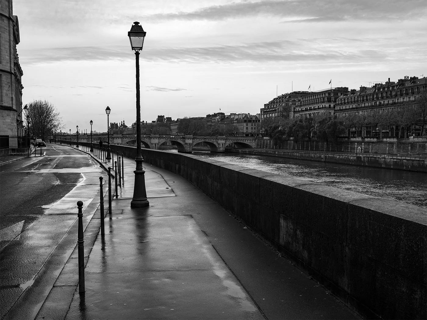 A black and white photo of a sidewalk along a river, with street lamps and a bridge in the distance, on a cloudy, possibly rainy day.