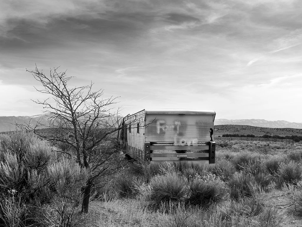 A desolate landscape with dry bushes, a leafless tree, and an abandoned camper in the distance.