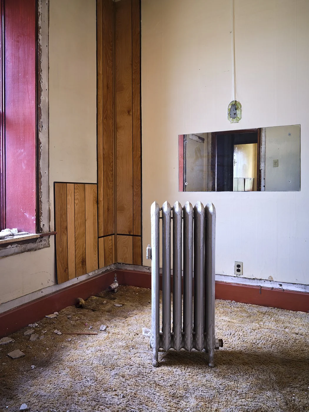 Room with beige walls, wooden and paneling accents, an old radiator, a mirror, a small window with red trim, and exposed drywall near the ceiling. The floor is covered in debris and carpet.