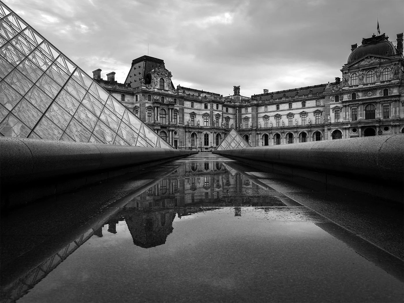 Black and white photo of the Louvre Museum in Paris, France, with modern glass pyramids in the foreground and the historic building in the background, reflected in a water feature.