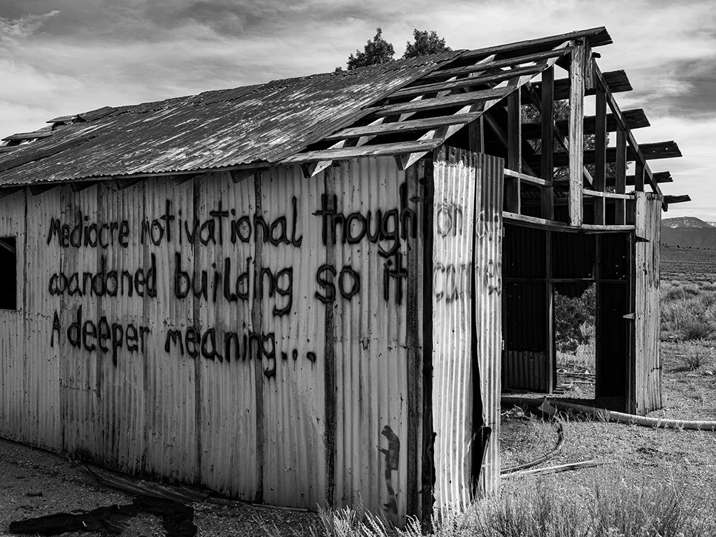 An abandoned, dilapidated metal building in a barren landscape with a motivational quote spray-painted on its side.