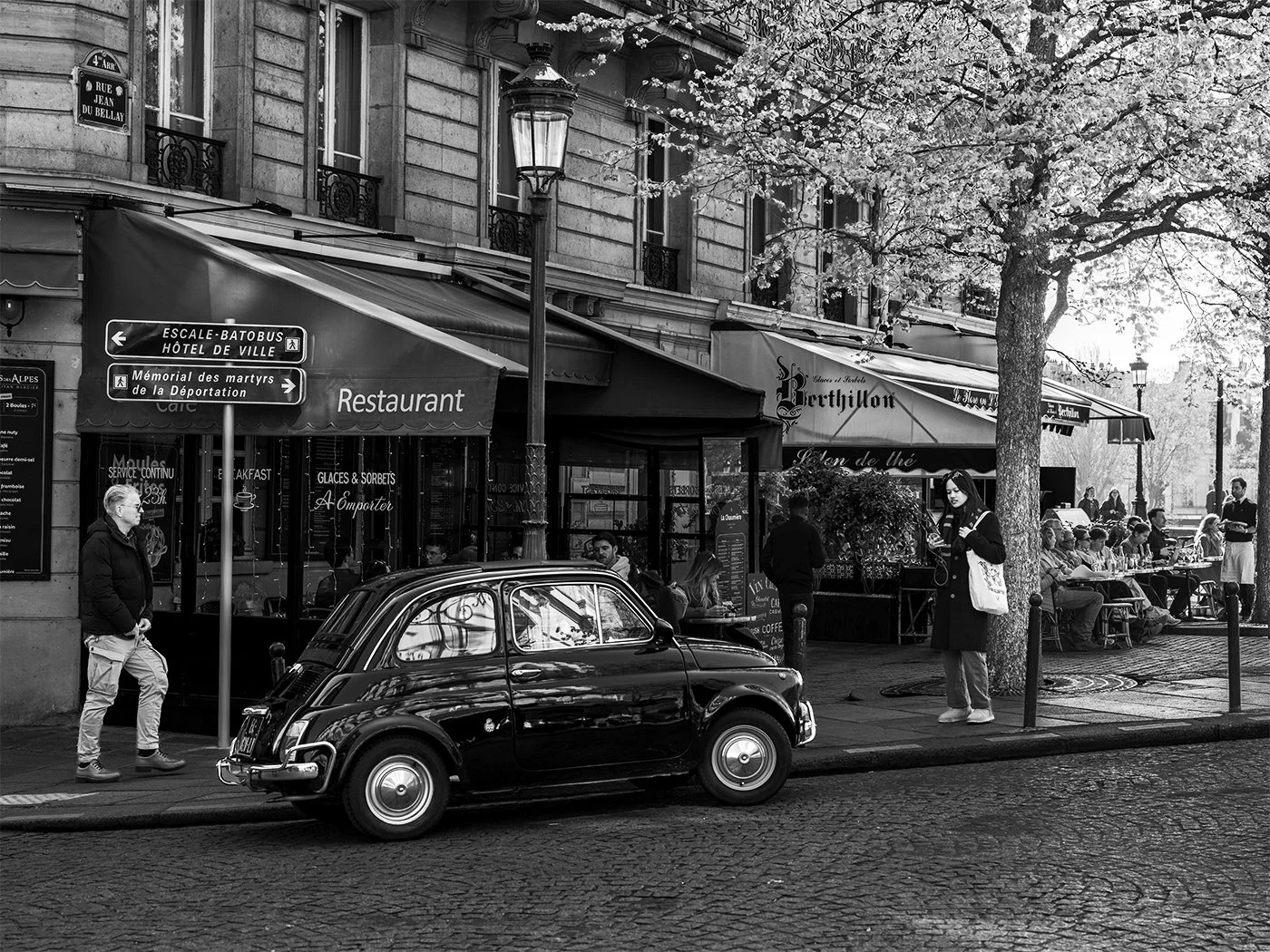 Street scene in Paris, France with a vintage black car on a cobblestone road, pedestrians, outdoor café seating under awnings, a large tree, and historic buildings in the background.
