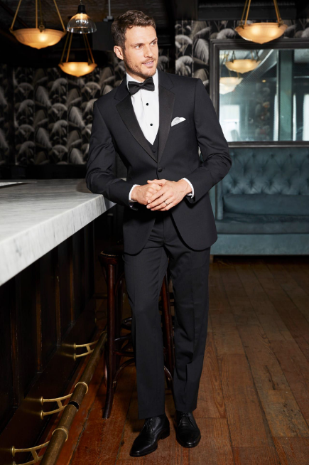 Man in black tuxedo leaning against a bar