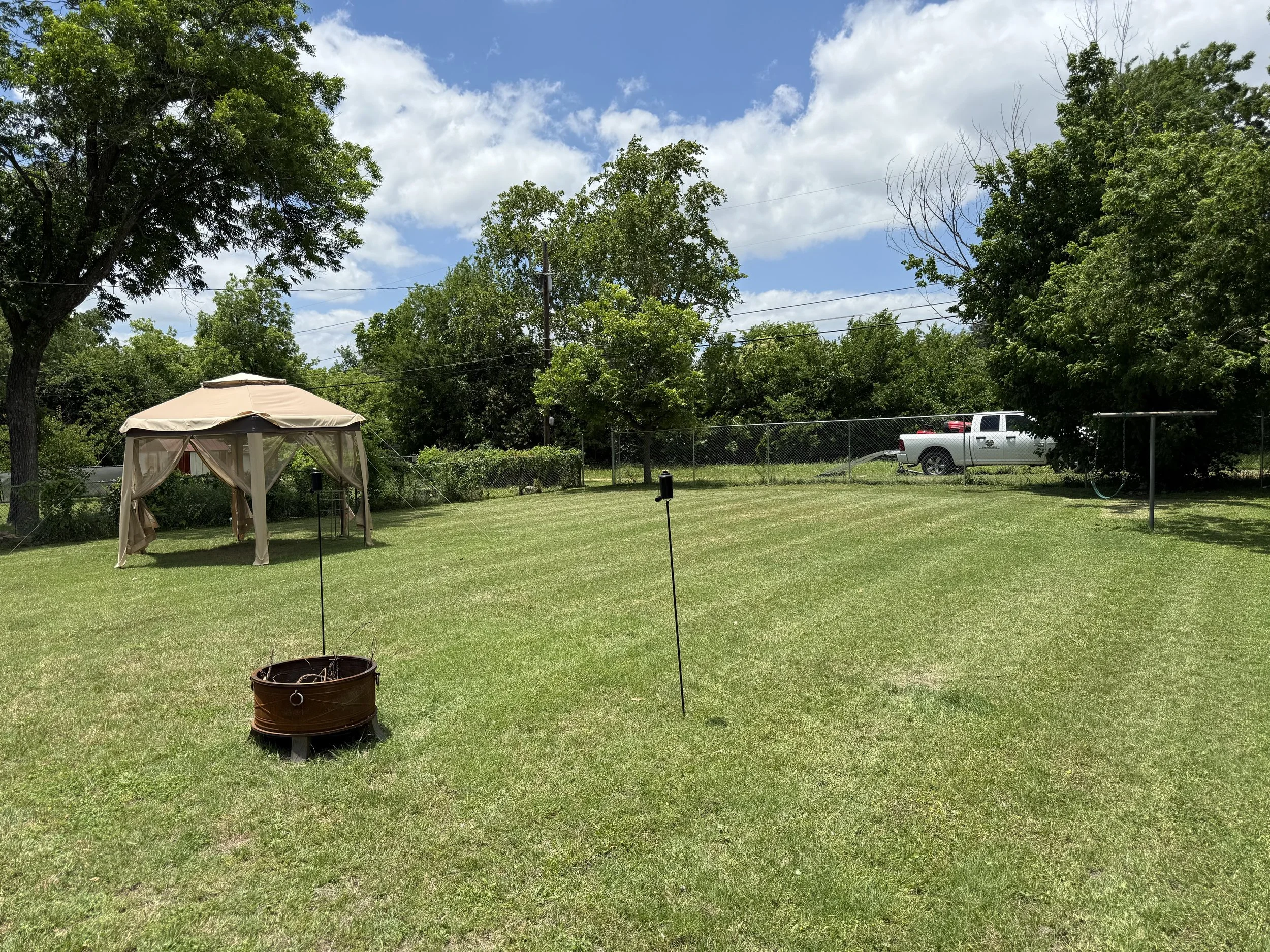 A backyard with trees, a gazebo with curtains, a firepit, a grassy lawn, a white pickup truck parked near a fence, and a blue sky with some clouds.
