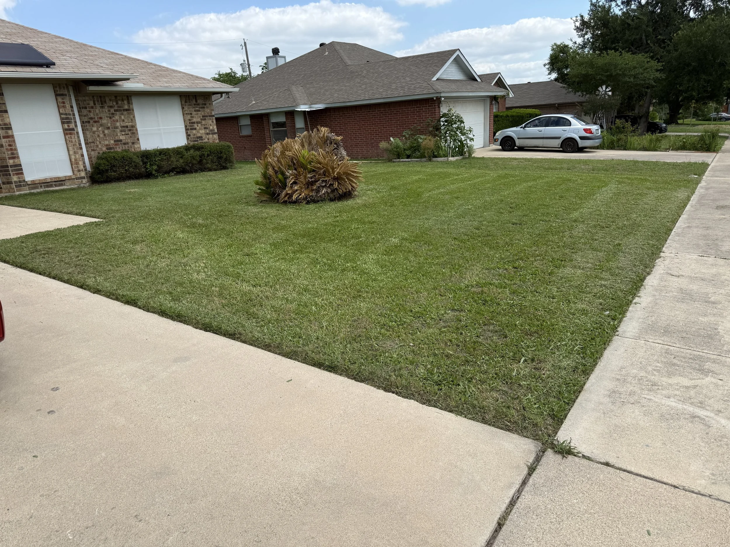 A suburban house with a brick exterior, small front yard, and a driveway with a silver car parked on it. The yard has green lawn, bushes, and a large plant. The sky is partly cloudy.
