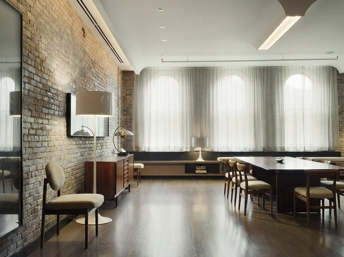 Modern dining room with brick wall, wooden floor, long table with chairs, and large windows with sheer curtains.