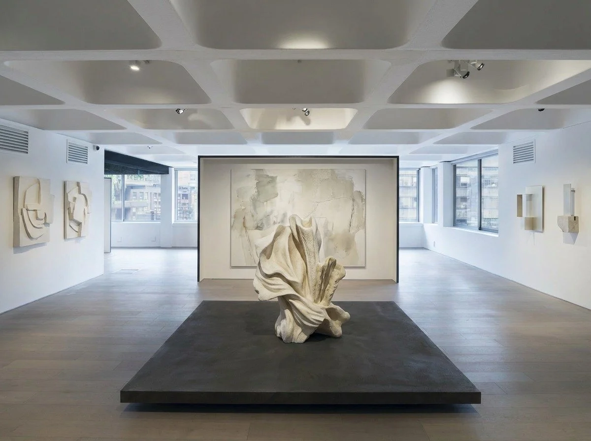 Modern art gallery with white walls and large windows, featuring sculptural and abstract artworks including a prominent white sculpted piece on a black platform.