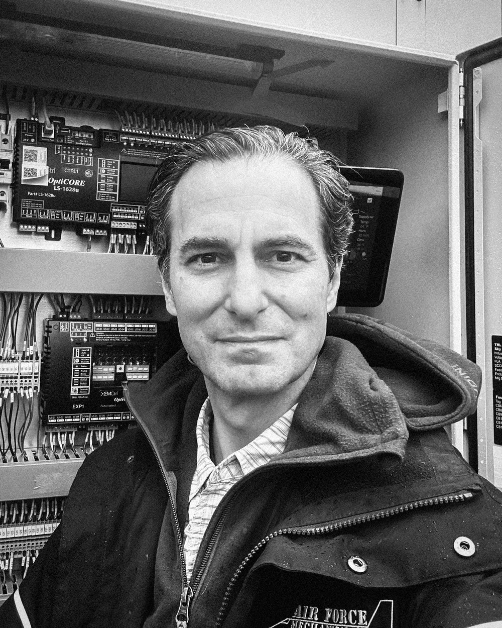 Kevin Kossi, founder of Air Force Mechanical Corp., in a jacket standing in front of electrical control panels.
