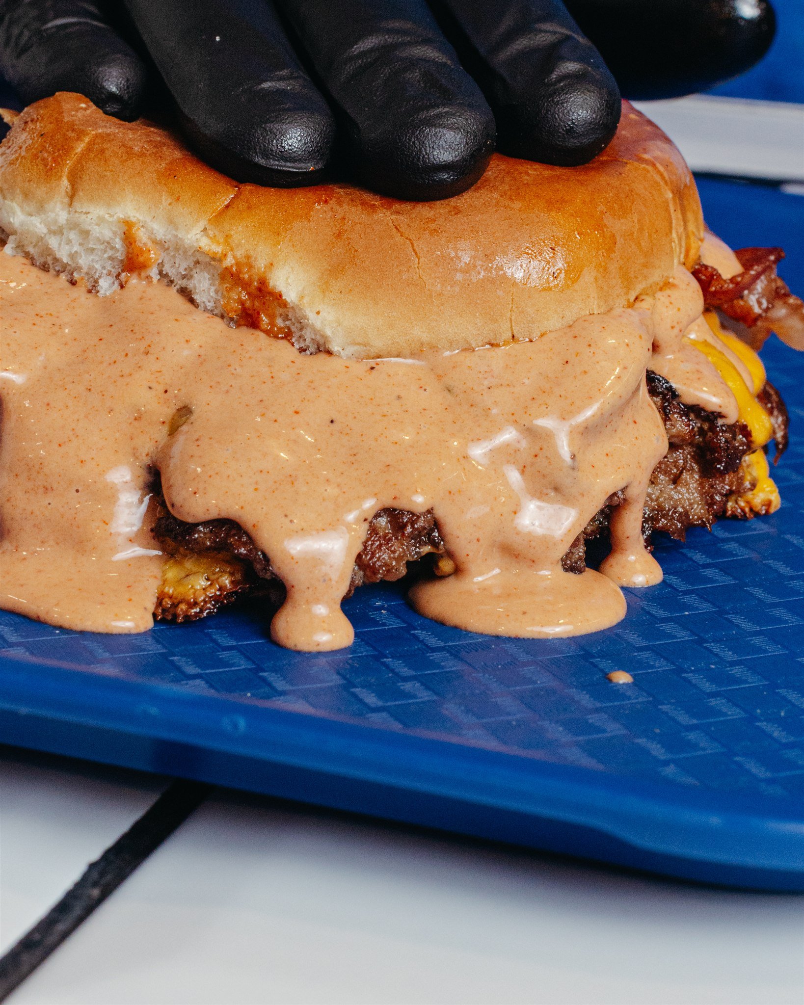 Close-up of a hamburger with melted cheese, bacon, and sauce on a blue plate, with a person's hand in a black glove resting on the bun.