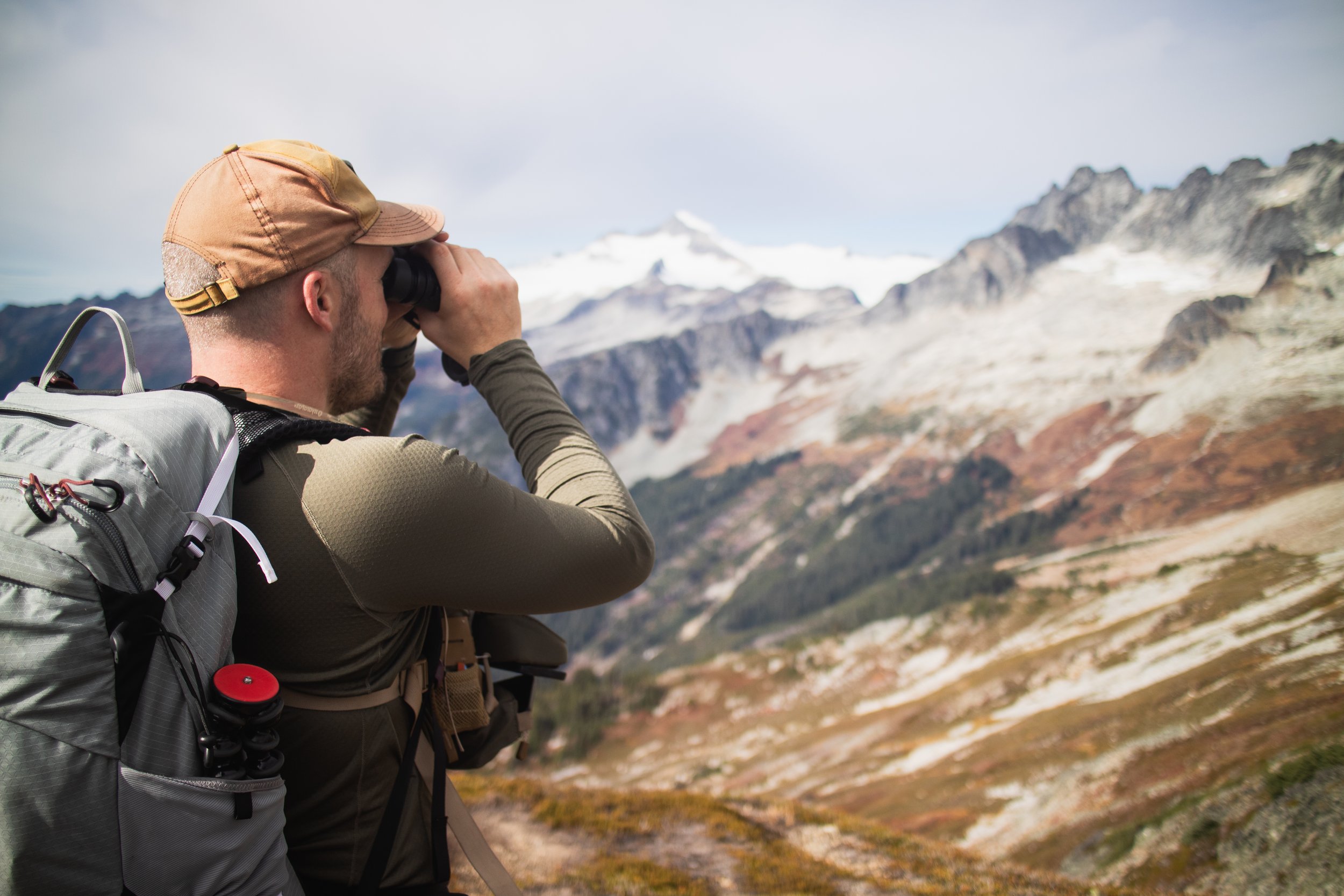 A man with light skin wearing a tan cap, gray backpack, and long-sleeve shirt, looking through binoculars at snow-capped mountains and rocky terrain in the distance on a cloudy day.