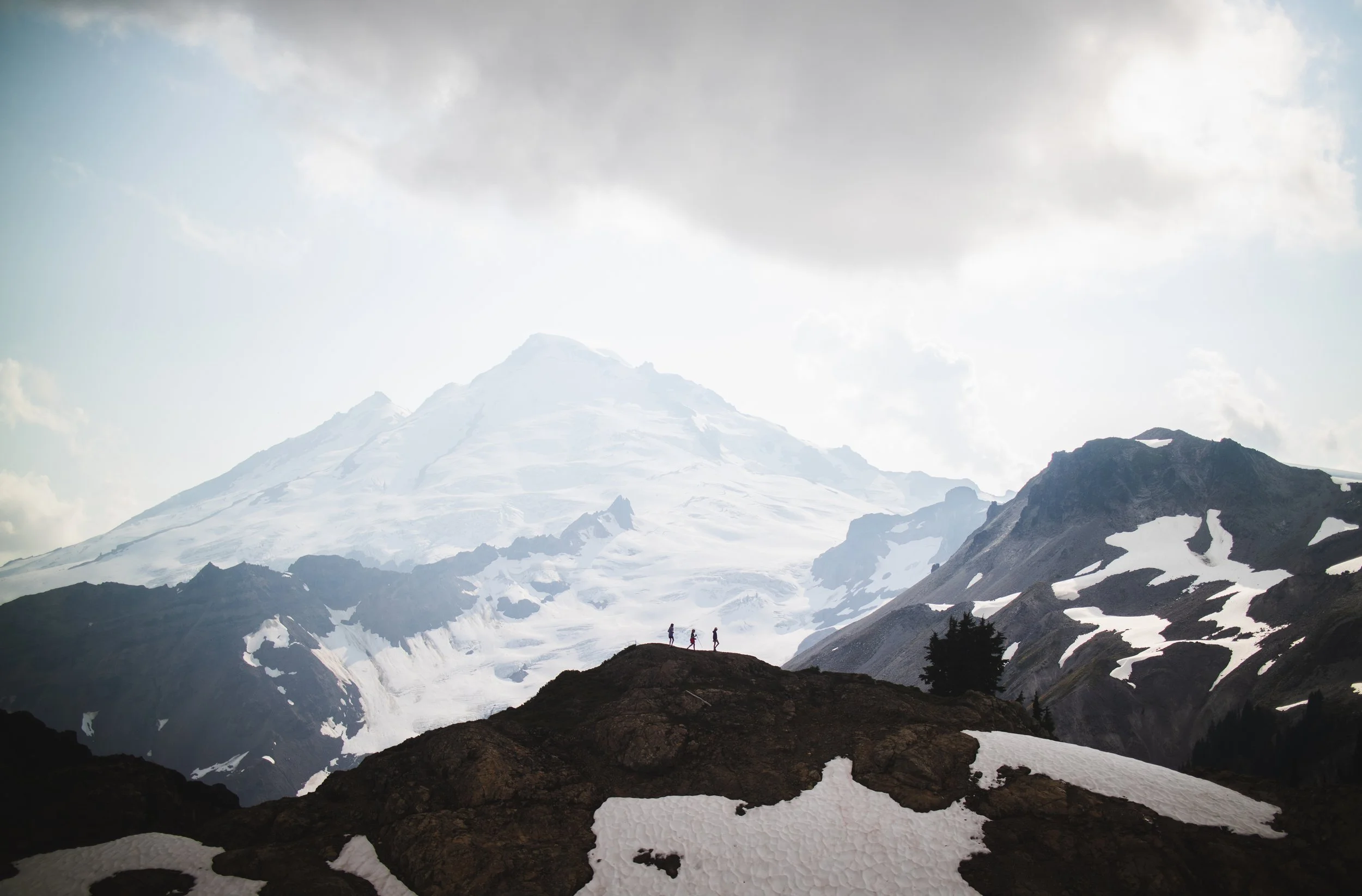 Snow-capped mountain with three hikers on a ridge in foreground, dark rocky slopes, and partly cloudy sky.