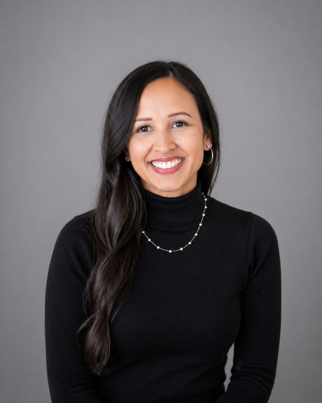 A smiling woman with long dark hair, wearing a black turtleneck sweater, a necklace, and hoop earrings, standing against a plain gray background.