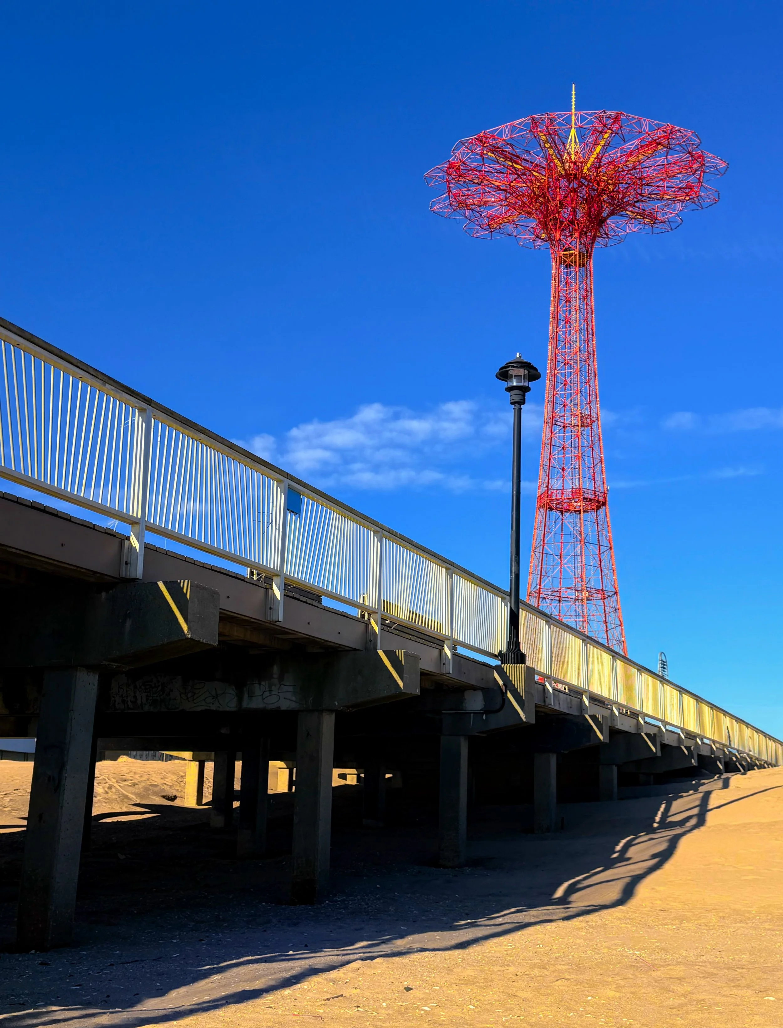 A red amusement park ride tower on a blue sky backdrop, with a pier and streetlamp in the foreground.