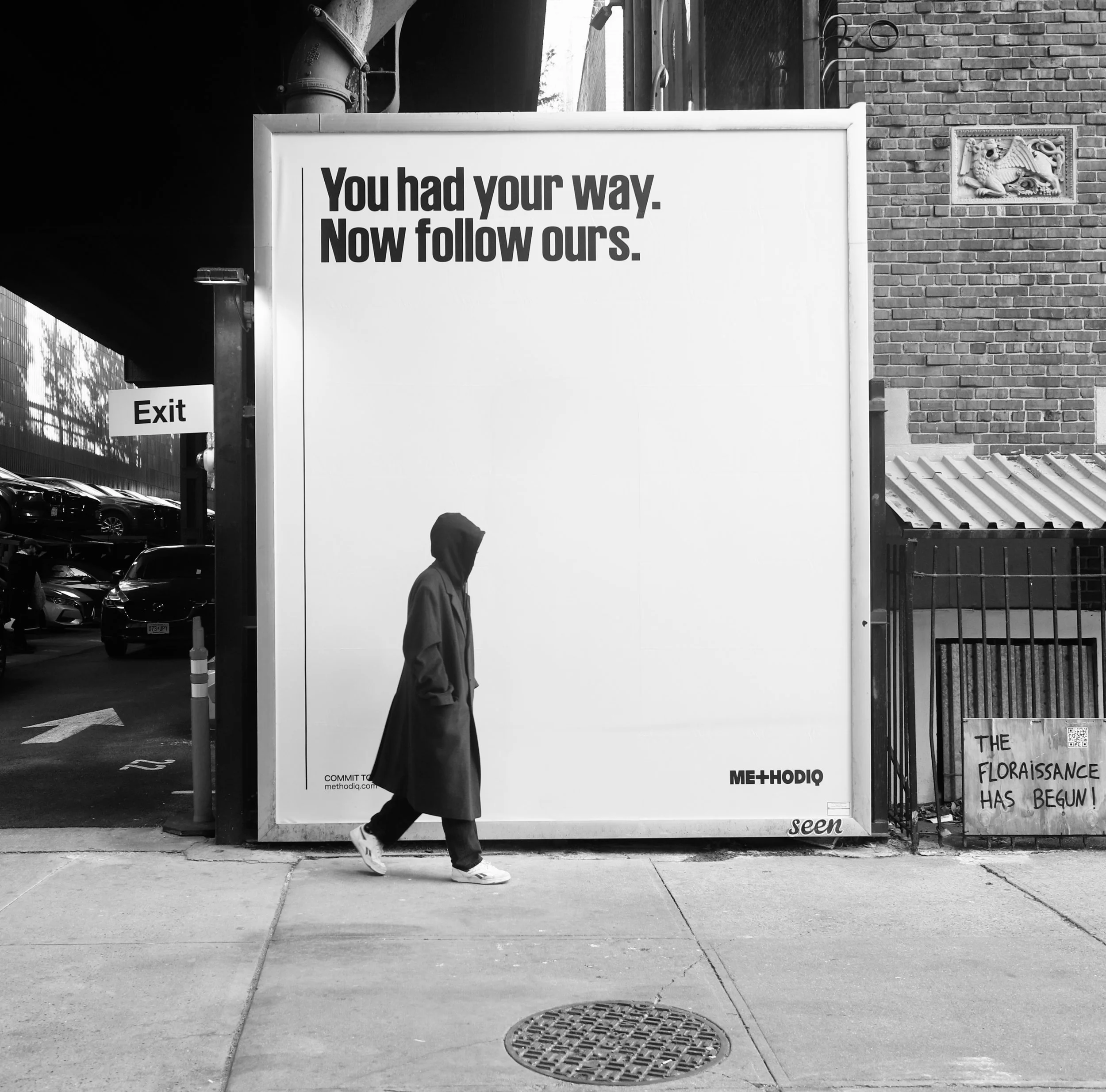 A person walking past a large sign with the text "You had your way. Now follow ours." in bold black letters. The sign appears to be at an outdoor location with parking and a brick building in the background. There is a smaller sign that says "Exit" a