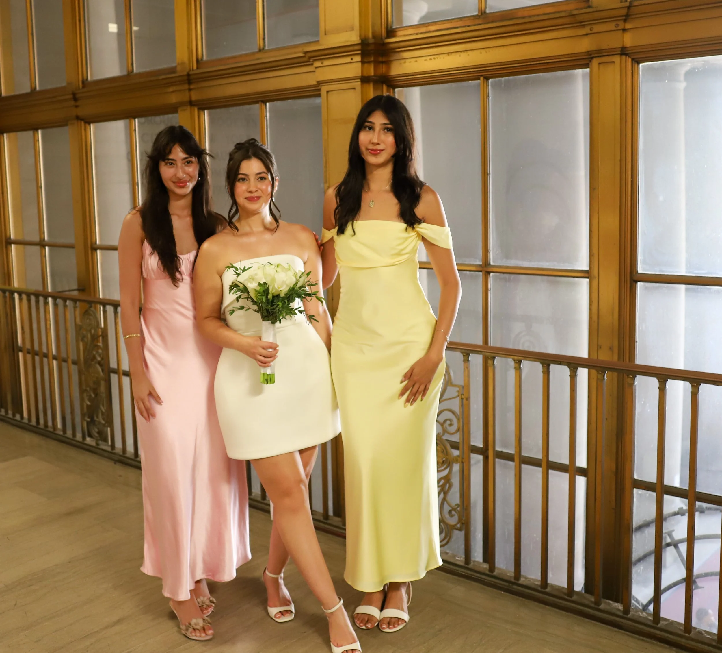 Three women in elegant dresses standing together indoors, with a golden railing and large windows in the background.