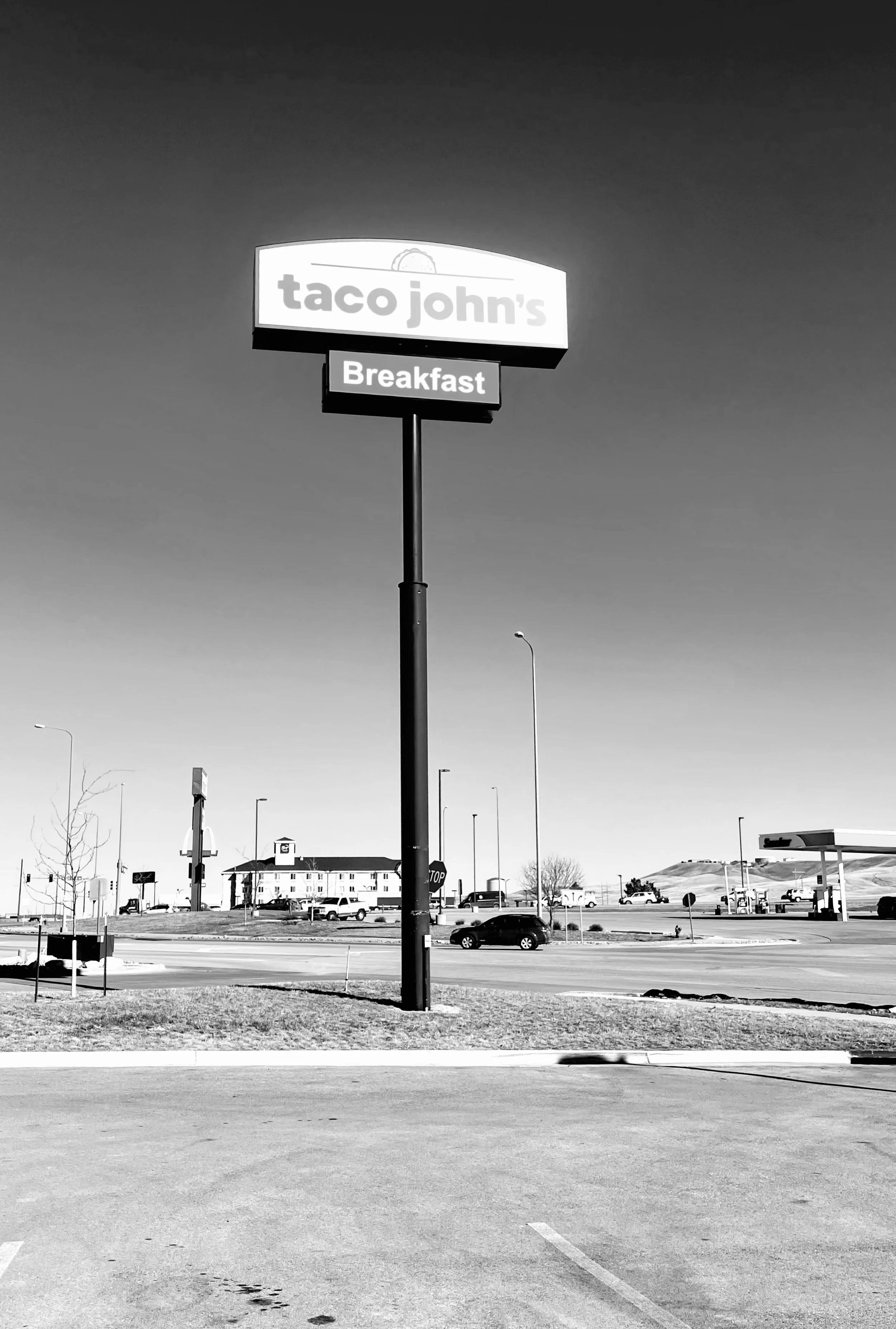 Black and white photo of Taco John's sign with a Taco icon, advertising breakfast, in a parking lot area with a few cars, buildings, and a gas station in the background.