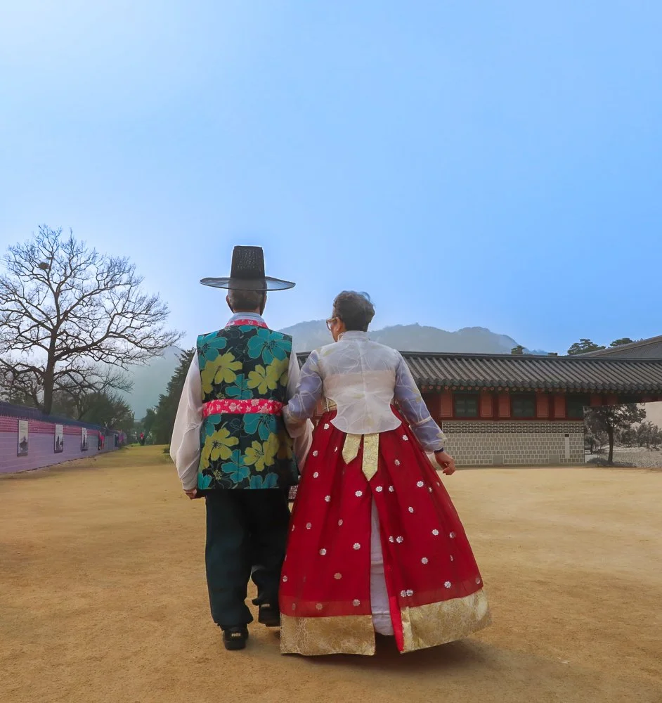 A man and woman dressed in traditional Korean hanbok, walking together outdoors near a traditional building with mountains in the background.