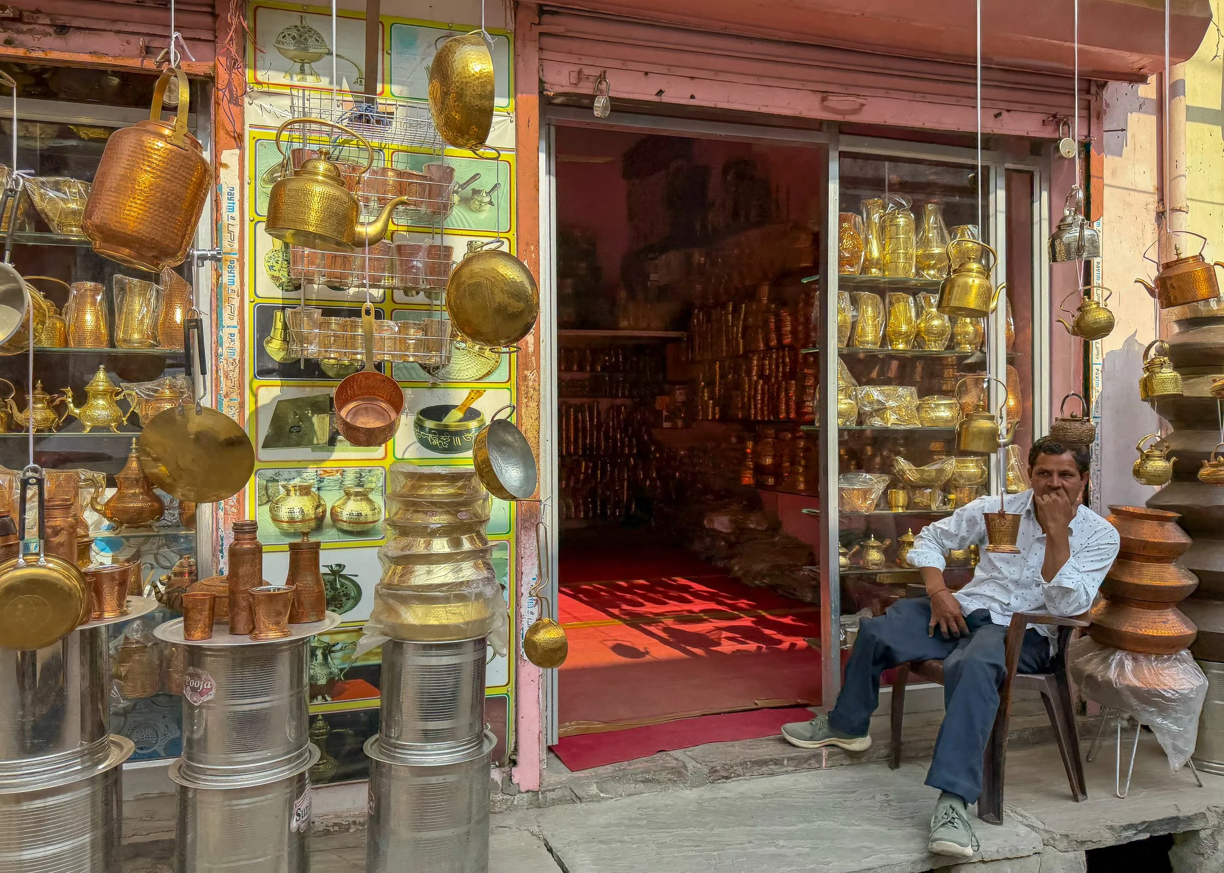 A man sitting outside a shop selling brass and copperware, with various pots, pendants, and utensils on display, some hanging and some stacked on shelves and countertops.