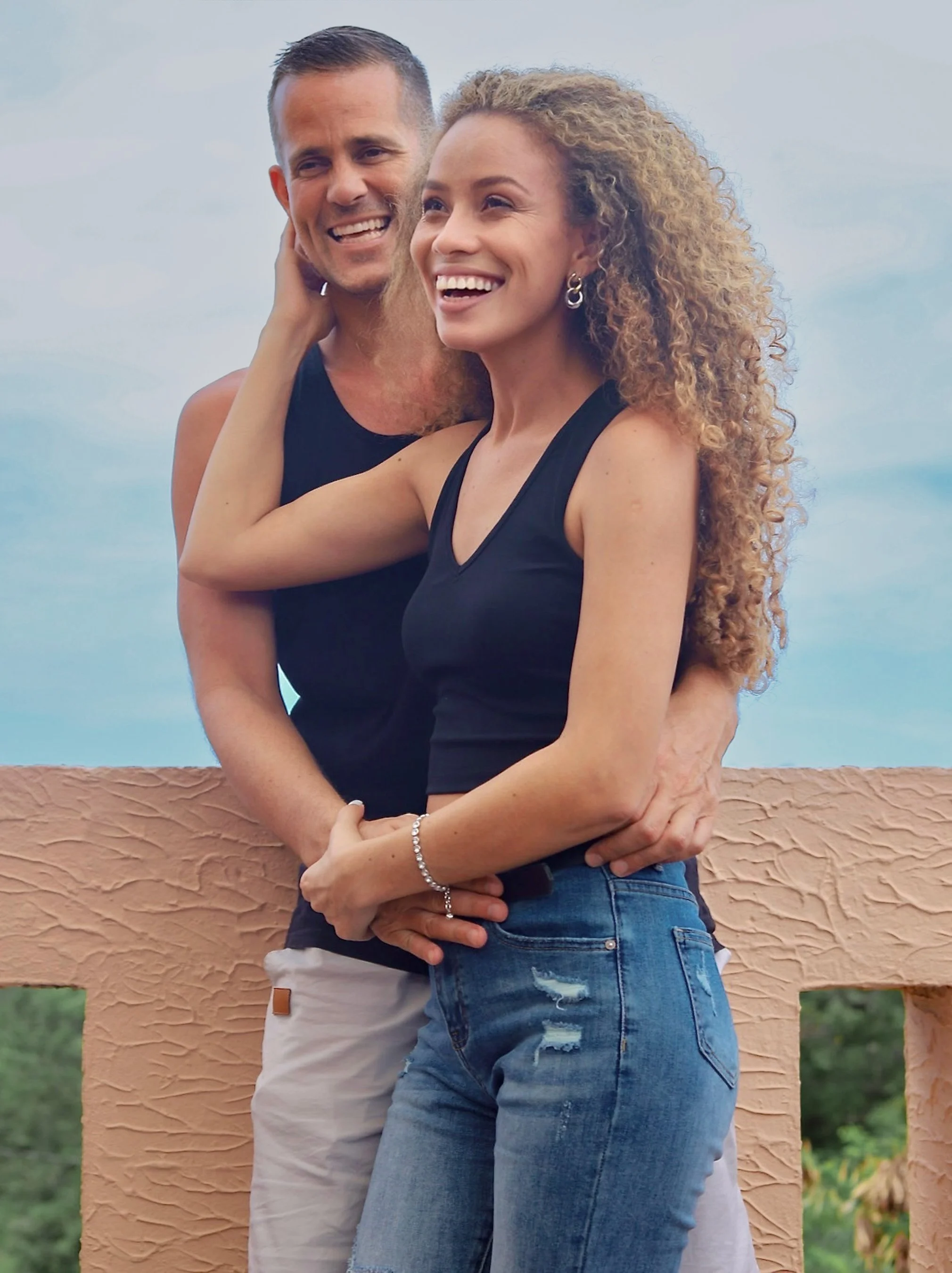 A happy couple standing outdoors, embracing each other with a clear blue sky and greenery in the background.