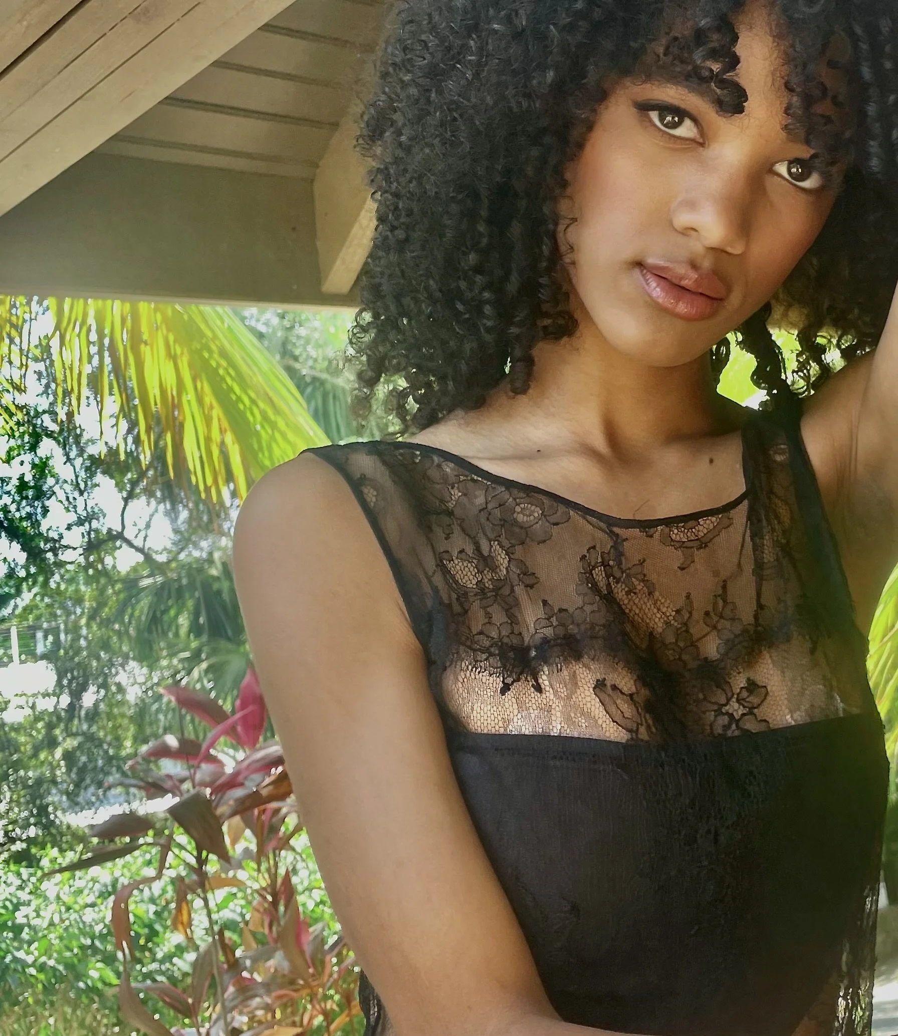 A woman with curly black hair and light brown skin wearing a black lace top, standing outdoors with greenery and palm trees in the background. Roatan 