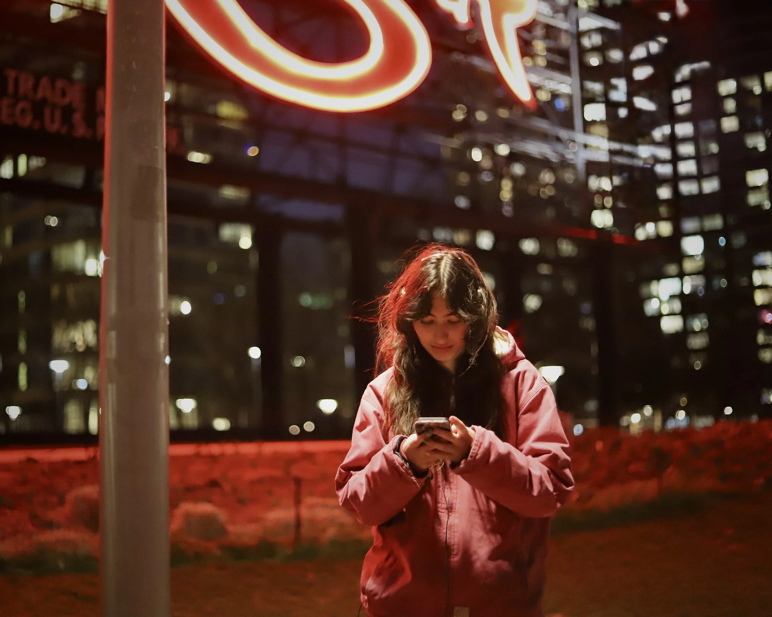 A young woman in a red jacket is standing outside at night, looking at her phone, with a city skyline in the background and neon lights above her.