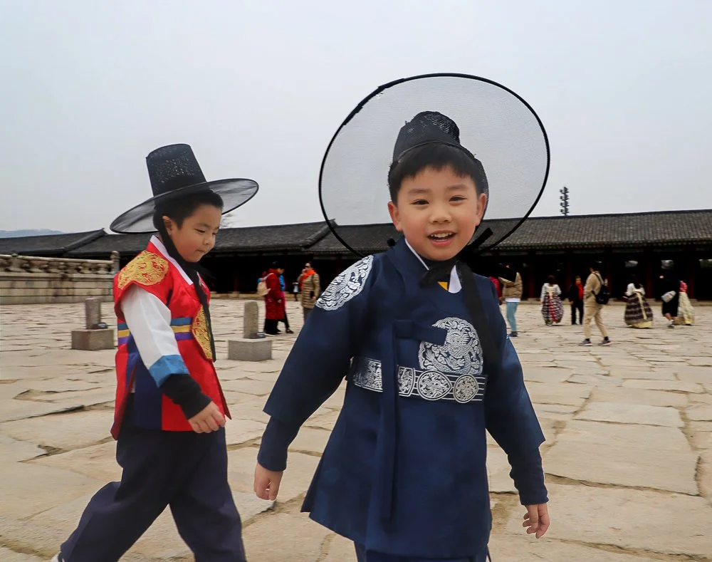 Two young boys dressed in traditional Korean hanbok clothing, with one boy smiling at the camera and wearing a black hat with a circular brim, at a historic site with stone pavement and other visitors in the background.