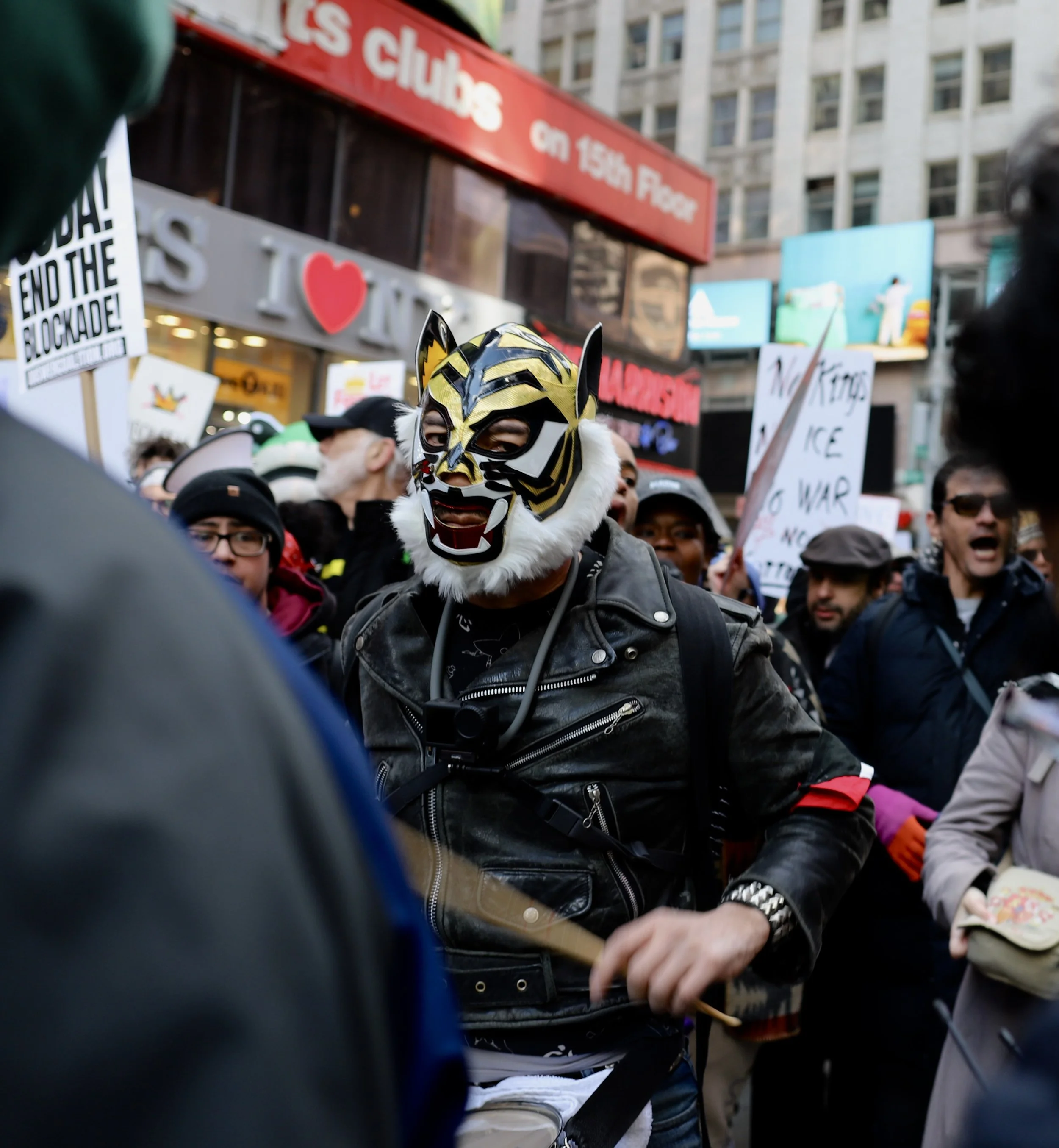 A person wearing a tiger mask and a black leather jacket participating in a protest or demonstration in a busy city square, surrounded by people holding signs.