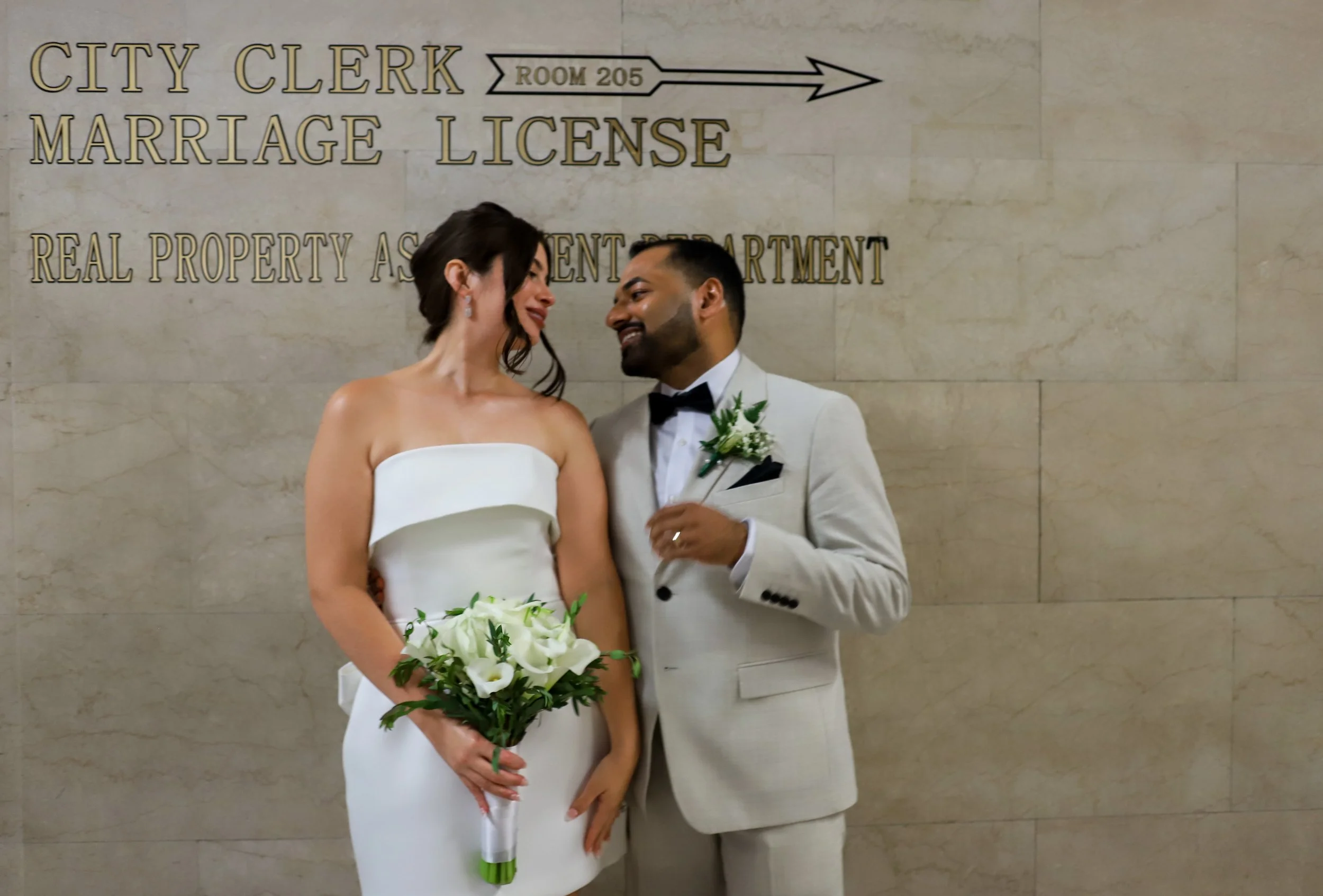 A newlywed couple, the bride in a white strapless dress holding a bouquet of white calla lilies, and the groom in a light-colored suit with a black bow tie and boutonniere, sharing a moment together in front of a sign for the city clerk's office.