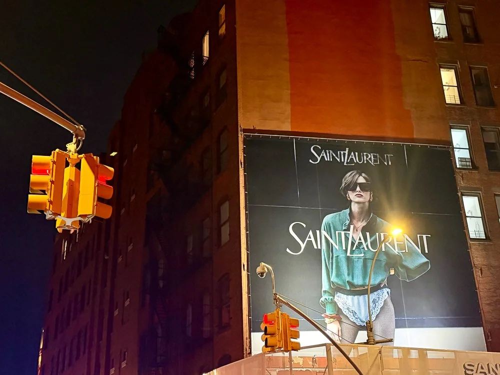 Nighttime city scene with a large billboard displaying a fashion advertisement for Yves Saint Laurent featuring a woman wearing sunglasses and a green jacket, with a traffic light hanging in the foreground.