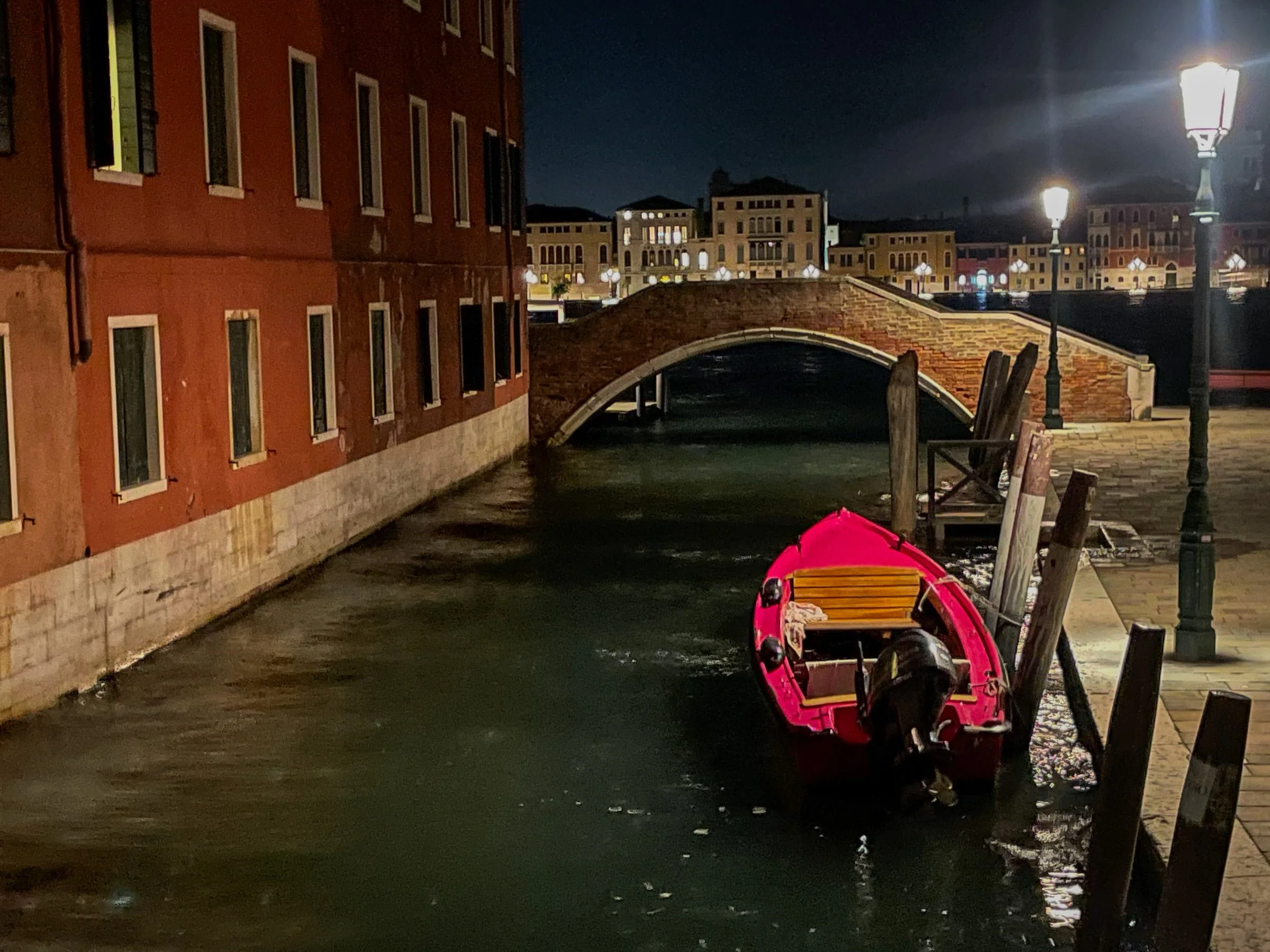 Nighttime scene of a small pink boat tied to a dock along a canal, with a stone bridge and illuminated buildings in the background.