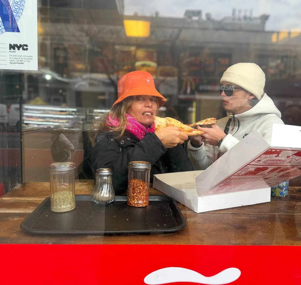 Two people sharing a slice of pizza through a restaurant window, with condiments on the table in front of them.