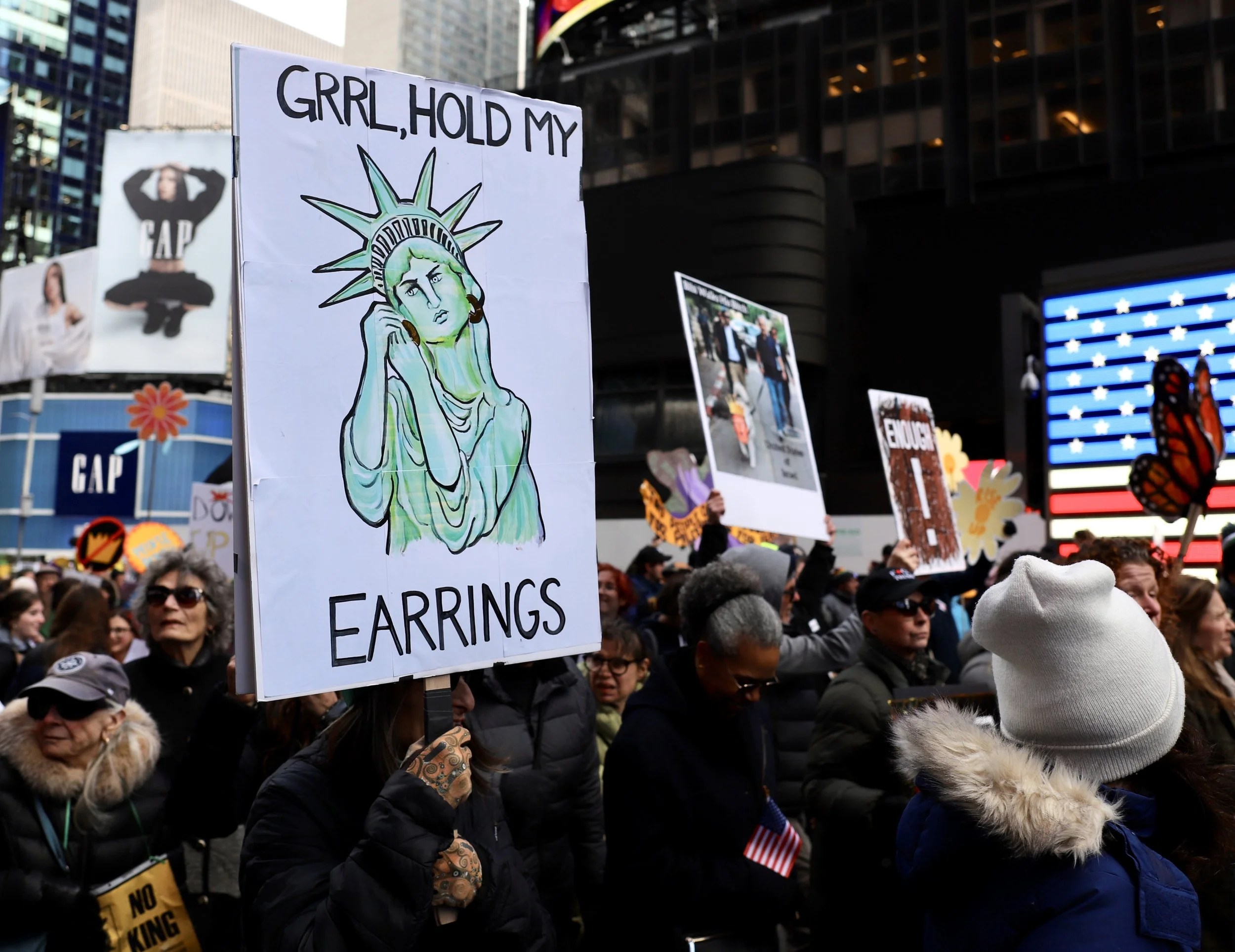 Protest crowd holding signs, including one with a drawing of the Statue of Liberty and the words "GIRL, HOLD MY EARRINGS" during a rally in an urban area with tall buildings and digital displays.